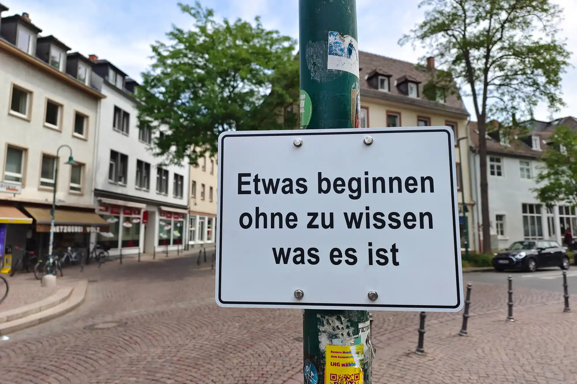 A sticker-covered white sign on a green pole above brown cobblestones in Darmstadt, framed by green trees in soft light.