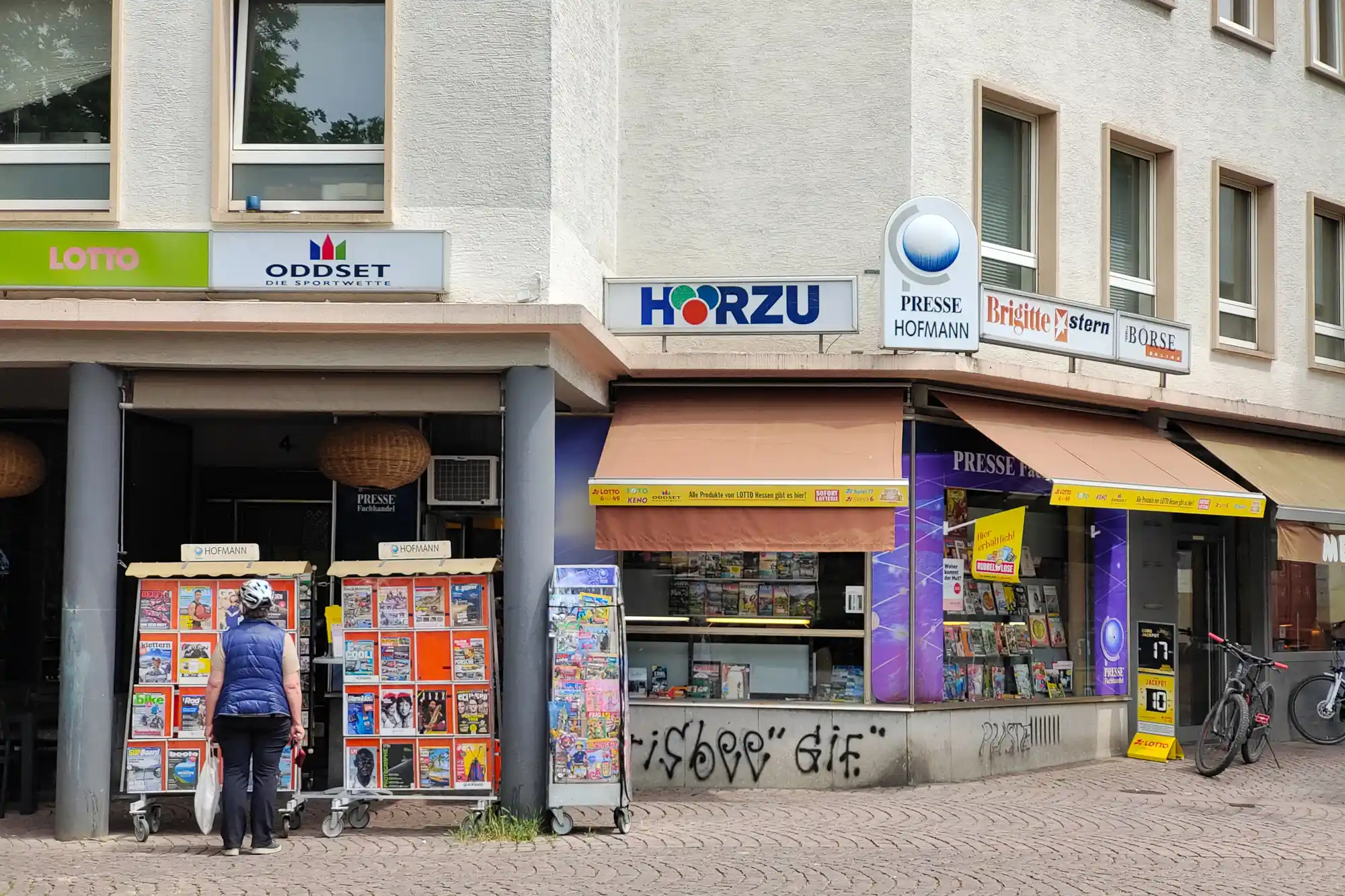A person in a blue vest standing by orange racks in the shade of tan awnings in Darmstadt, on a summer day.