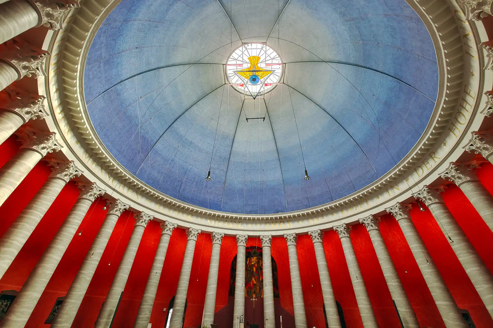 Looking up into a blue dome with a yellow stained-glass center in Darmstadt, framed by red walls and white columns.