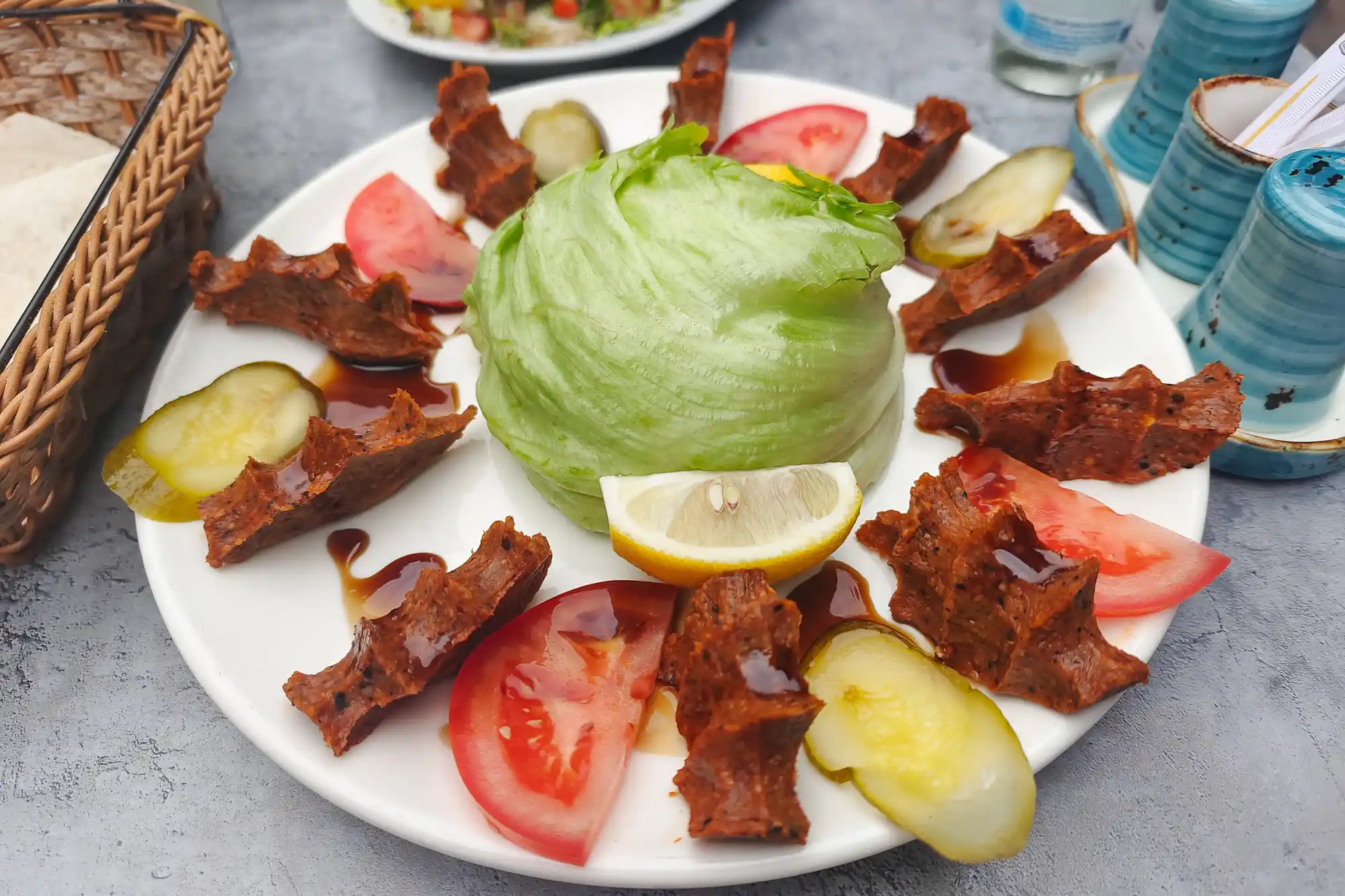 A salad of green lettuce with brown pieces and red slices on a white plate in Darmstadt, beside blue cups and a woven basket.