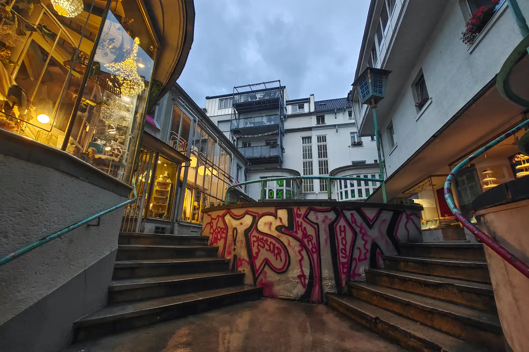 Two staircases framing a pink and black graffiti wall in Darmstadt, with warm yellow light glowing from a glass shopfront.