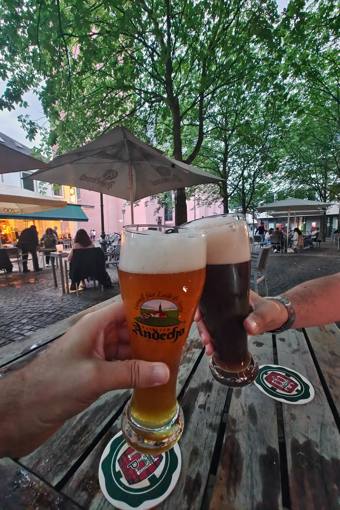 Two hands holding an amber and a dark beer over a wooden table on a summer evening in Darmstadt, with green trees and a grey umbrella.