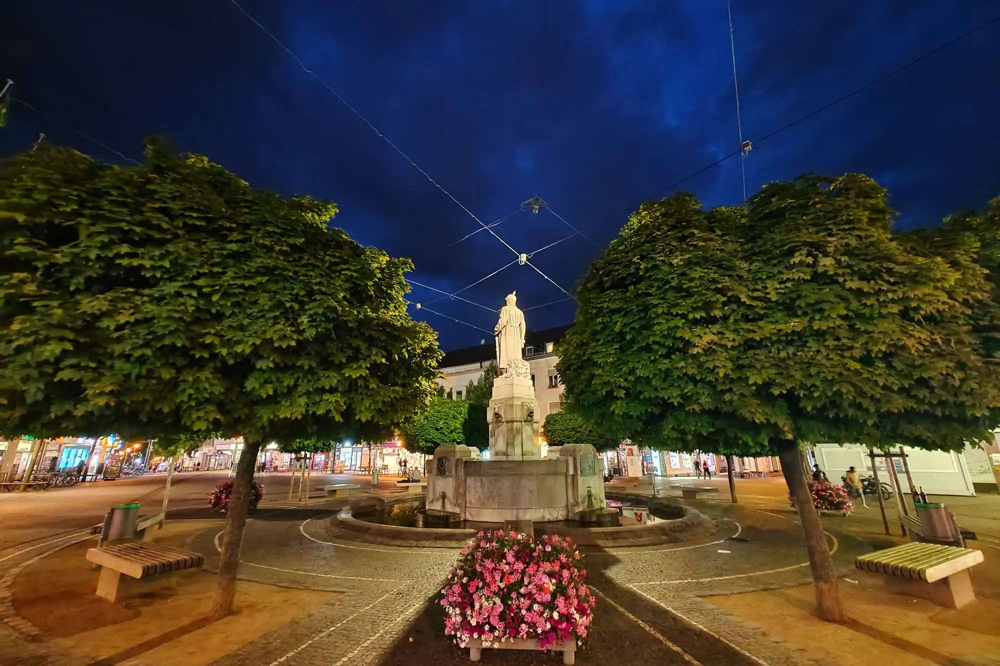 A stone statue standing at night under a deep blue sky in Darmstadt, framed by pink flowers and green trees.