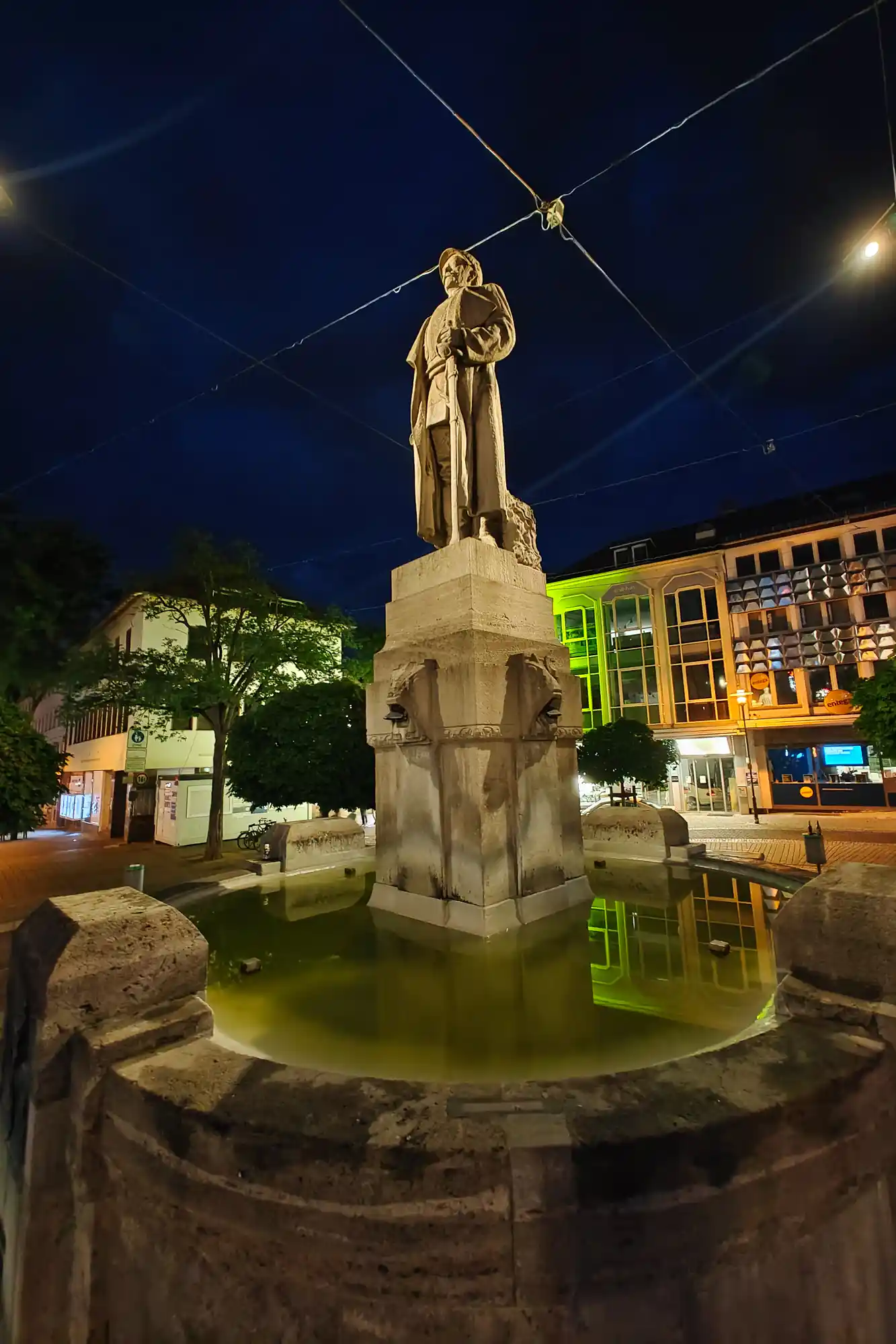 A stone statue standing in a basin of green water in Darmstadt, with overhead wires crossing the dark blue night sky.