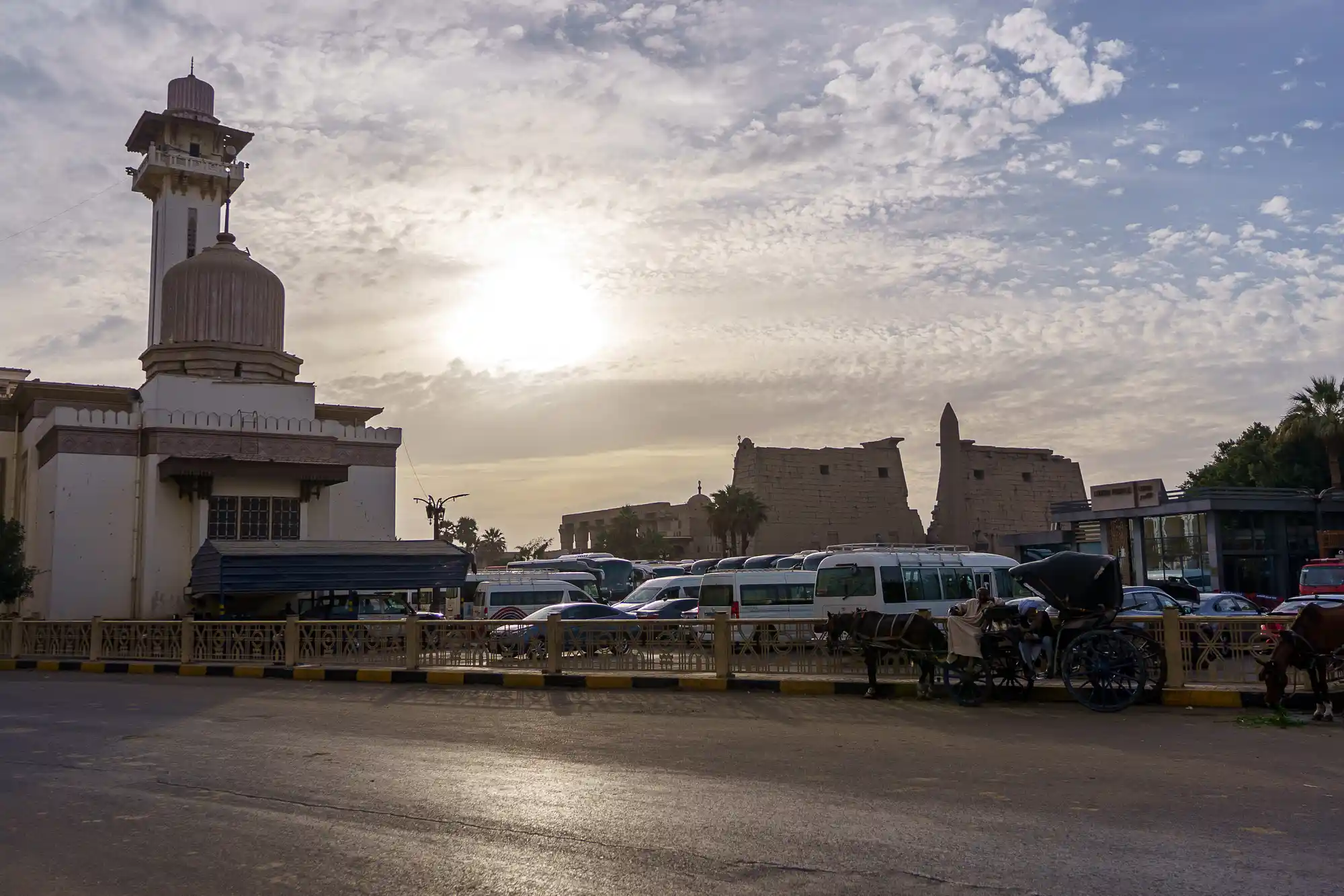 A white domed building with a tower in low sunlight in Luxor, with horse carriages and white vans lining the paved square.