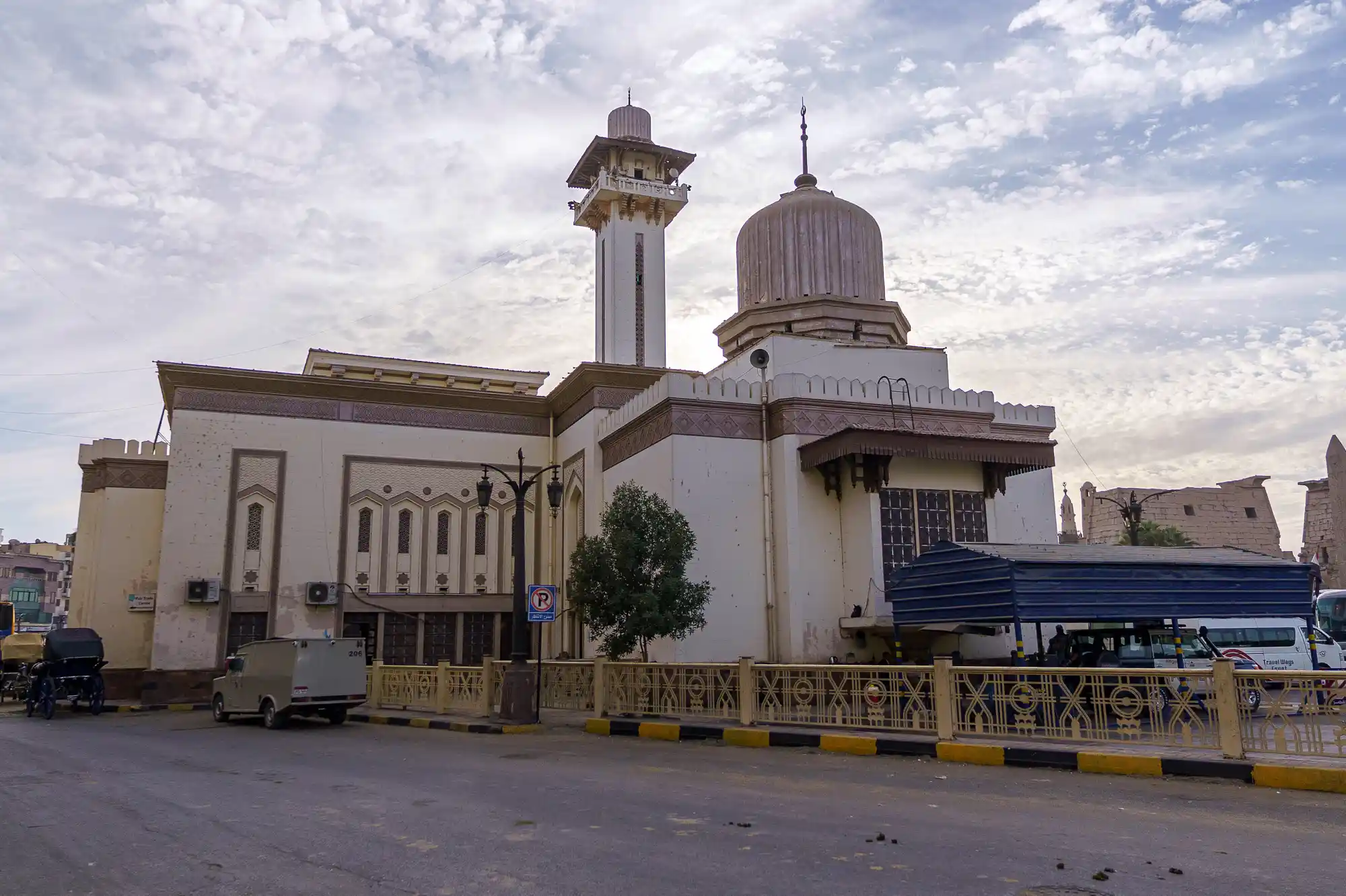 A white building with a ribbed dome and tower under a cloudy Luxor sky, with a yellow railing and a tree nearby.