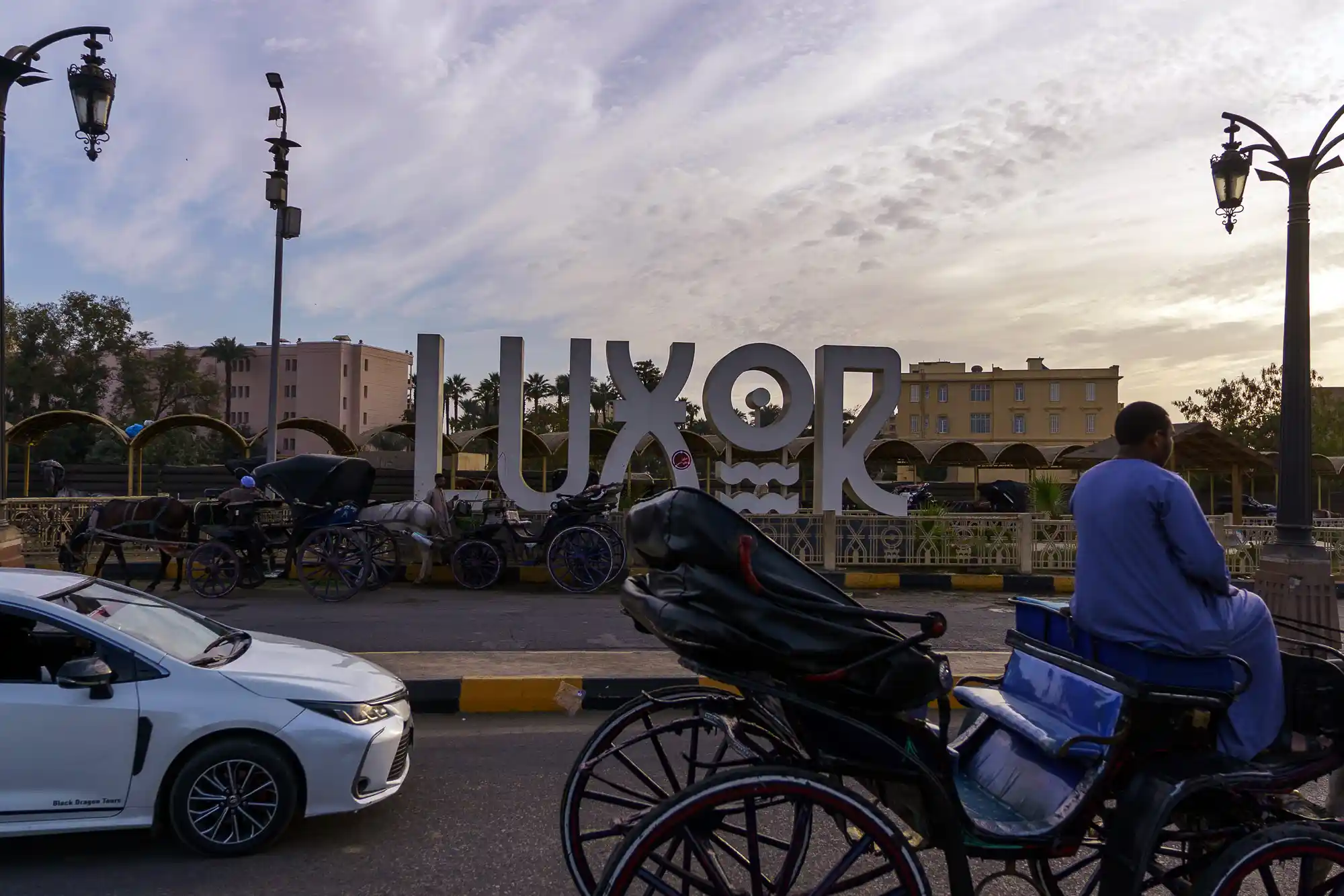 A person in blue sits on a black horse carriage with blue padding in Luxor, beside a white car and large white letters.