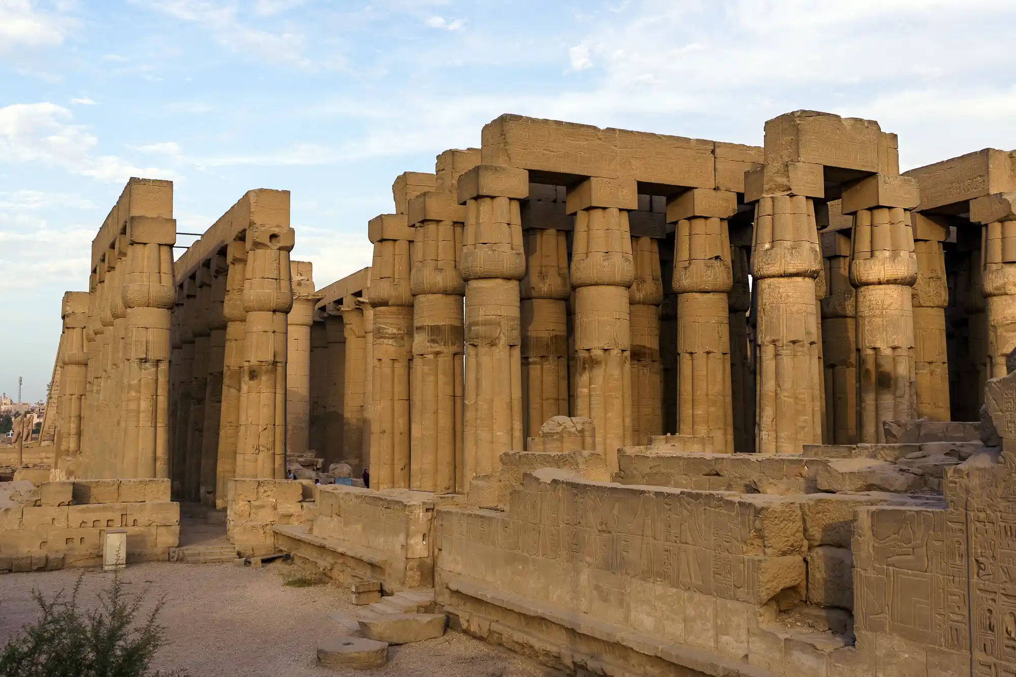 Carved sandstone columns of the Luxor Temple in warm light, framed by a pale blue sky and sandy ground.