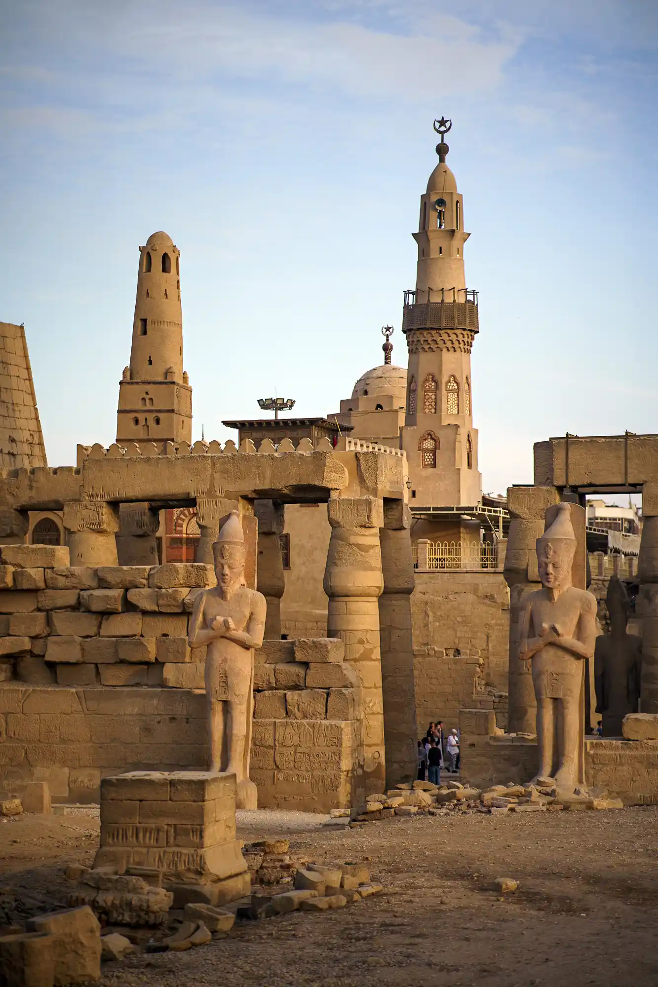 Two stone statues at the Luxor Temple entrance in warm light, with visitors walking among the sandstone blocks.