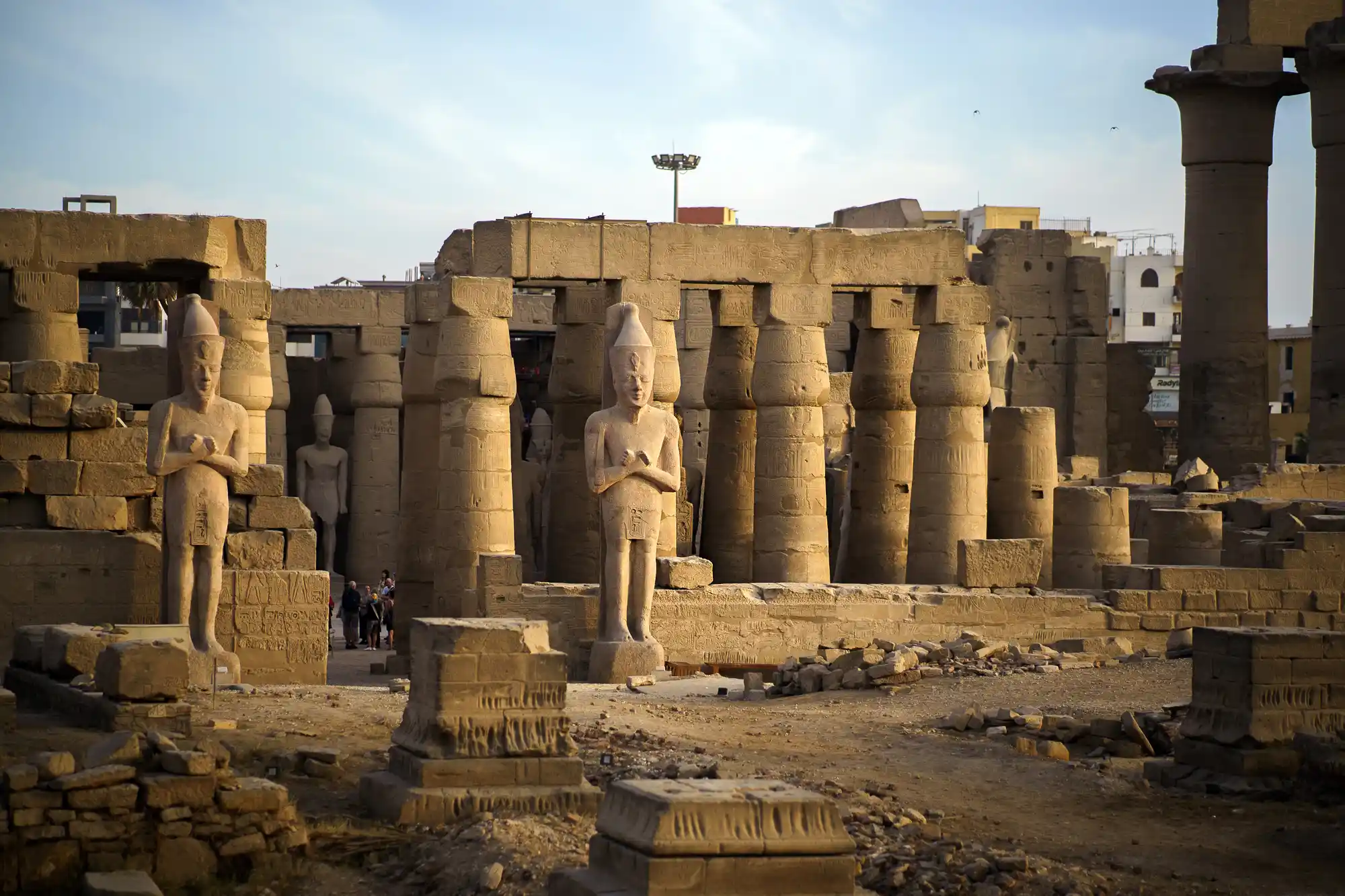 Stone statues flanked by columns at the Luxor Temple, with a few visitors walking on the sandy ground below.