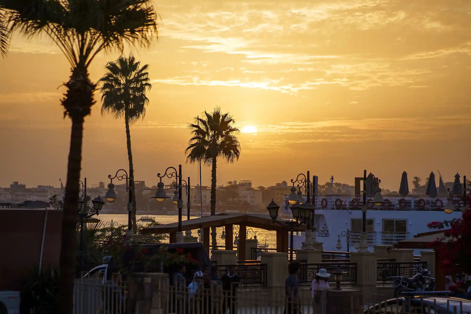 Silhouetted palm trees against golden sunset light on the Nile promenade in Luxor, with people near ornate lamps and a white boat.