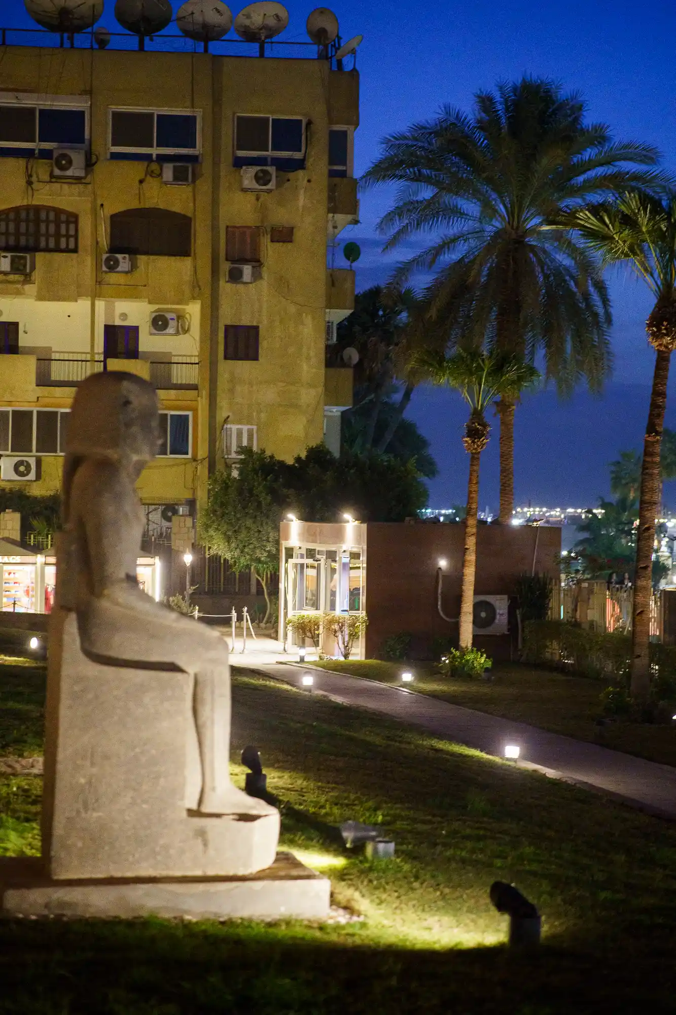 A stone statue in fading light in Luxor, with tall palms and a yellow building with satellite dishes behind it.