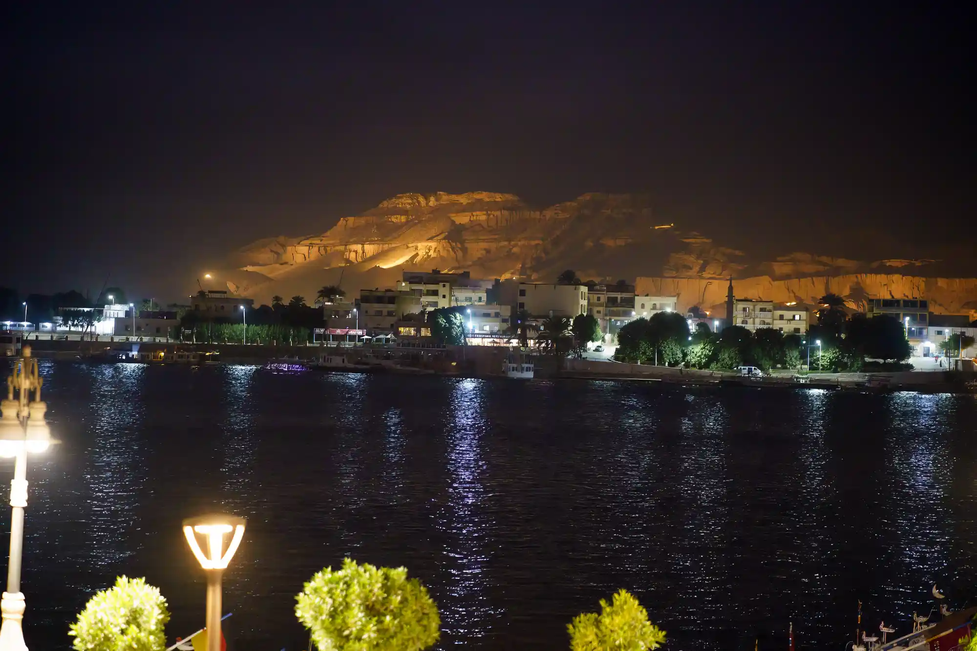 The Nile in Luxor at night, reflecting warm lights from illuminated buildings, with a golden-lit ridge on the far bank.