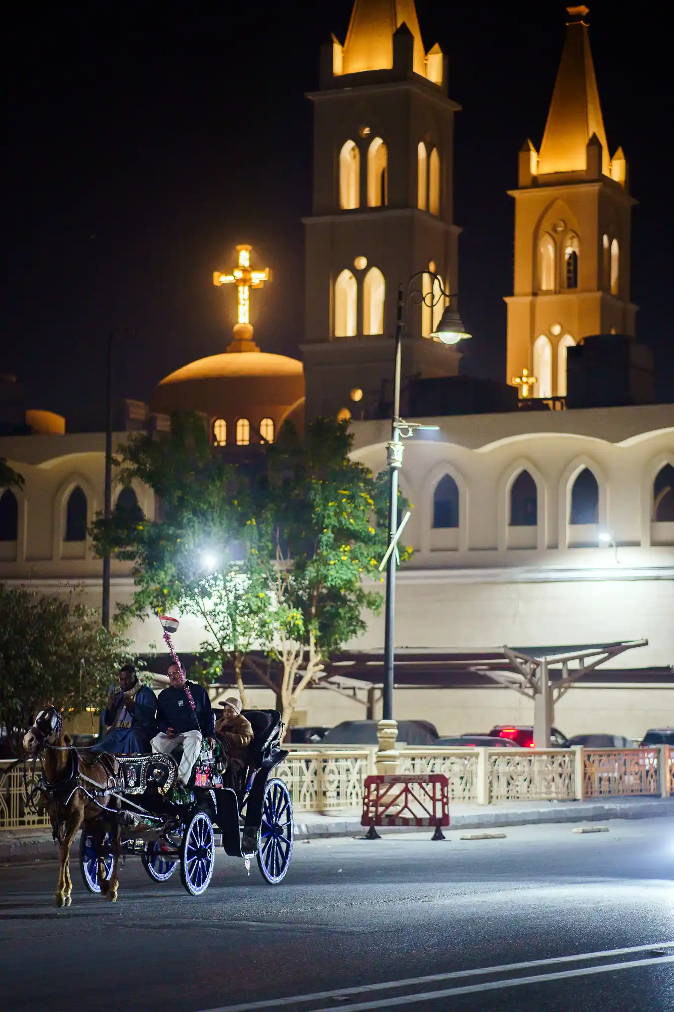 A horse-drawn carriage with three people in warm evening light in Luxor, passing a building with golden lighting and arched windows.