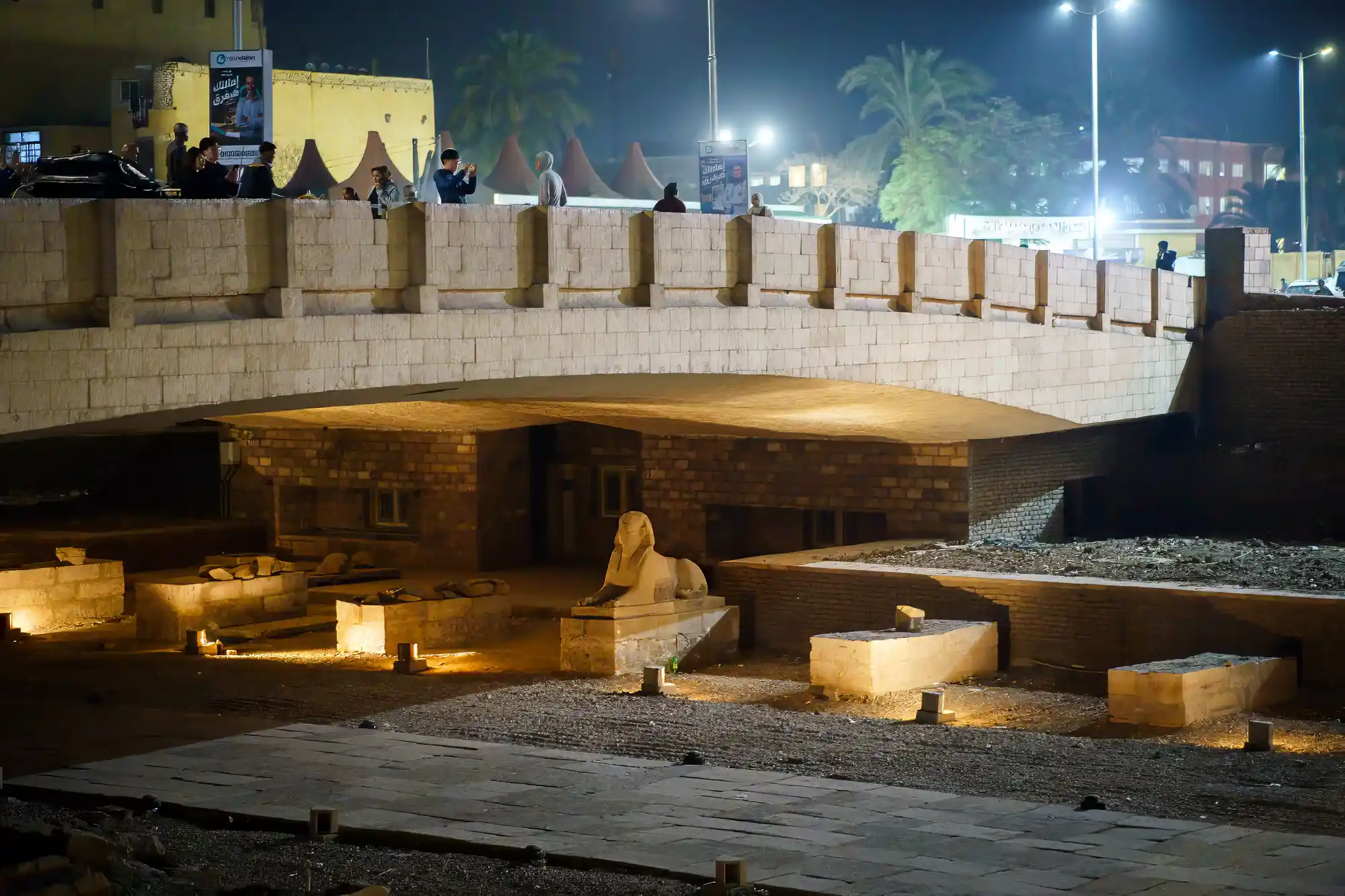 A stone sphinx under a lit arch in Luxor at night, with people standing on a tan stone bridge above.