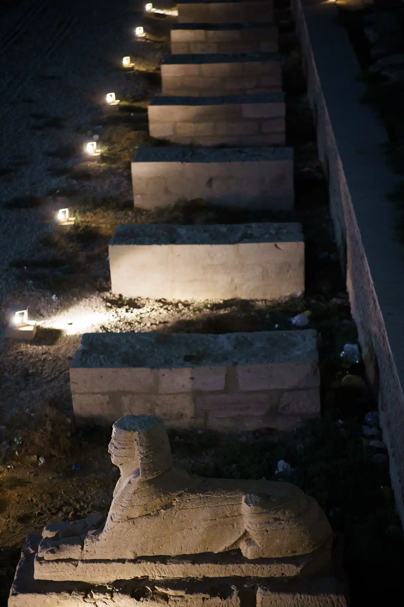 A stone sculpture on a block in Luxor at night, lit by warm yellow lights above the dark gravel ground.