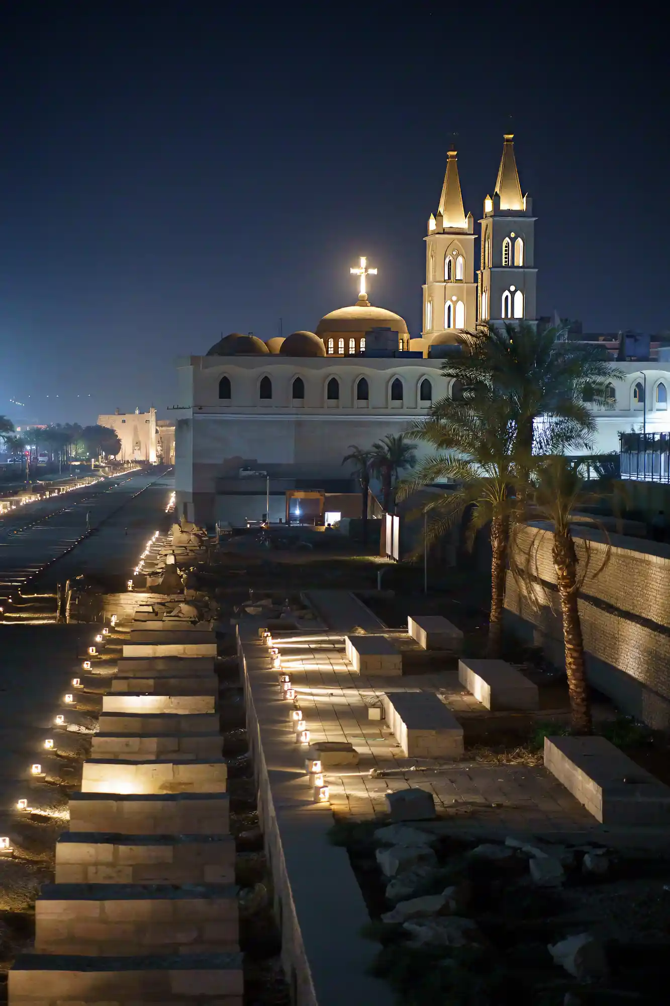 A church with golden domes and two towers lit up at night in Luxor, with small lights along stone paths and palm trees.