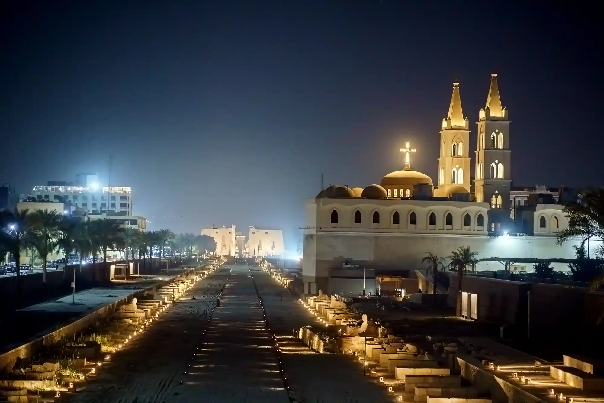 An illuminated church with two spires and a cross in Luxor at night, with warm yellow lights lining a wide path.