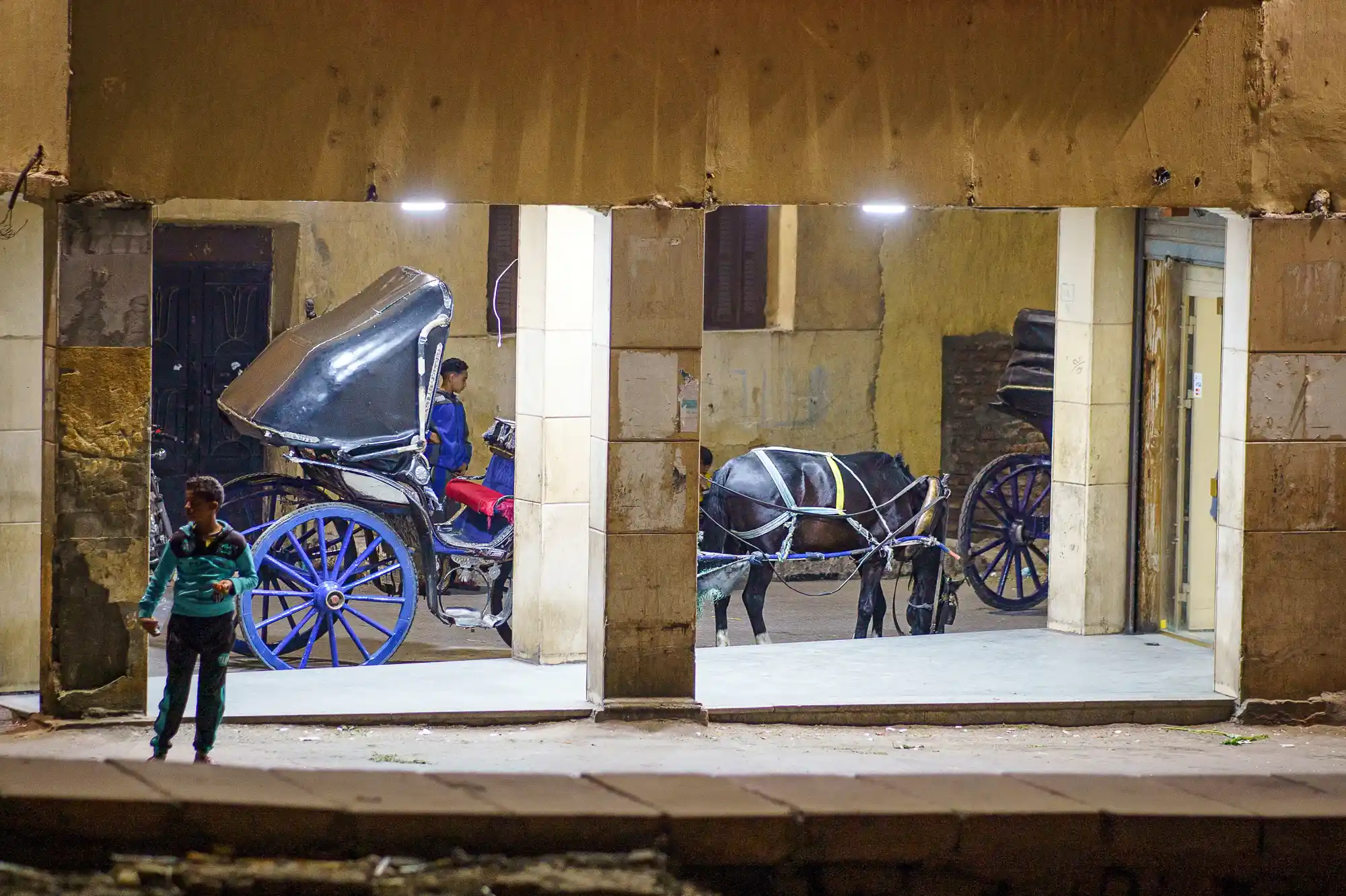 A horse-drawn carriage with blue wheels and a black top under street lights in Luxor, with a person in a green shirt nearby.