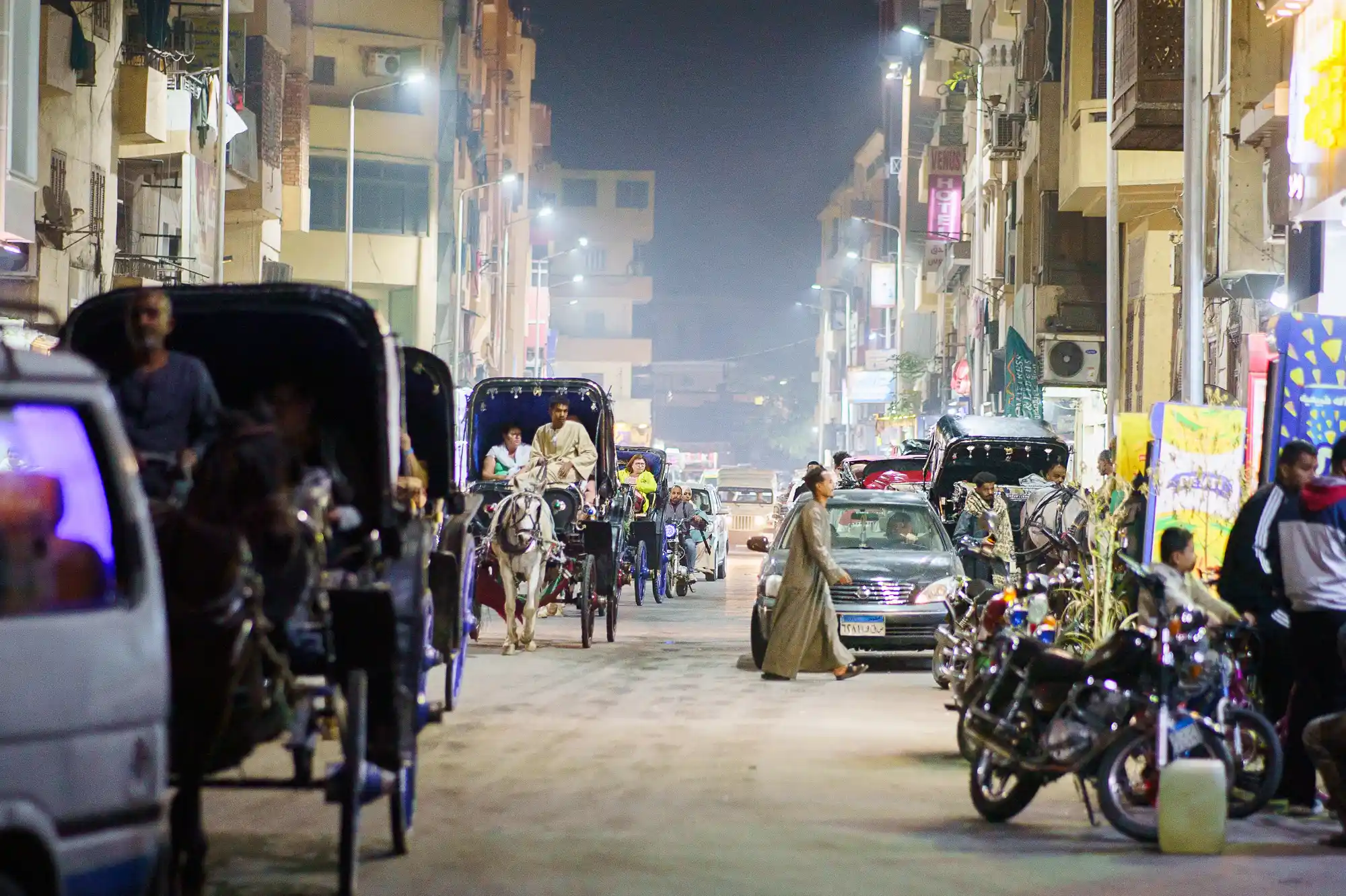 A horse carriage in a Luxor street at night under overhead lights, as a person in a long tan robe crosses past dark motorcycles.
