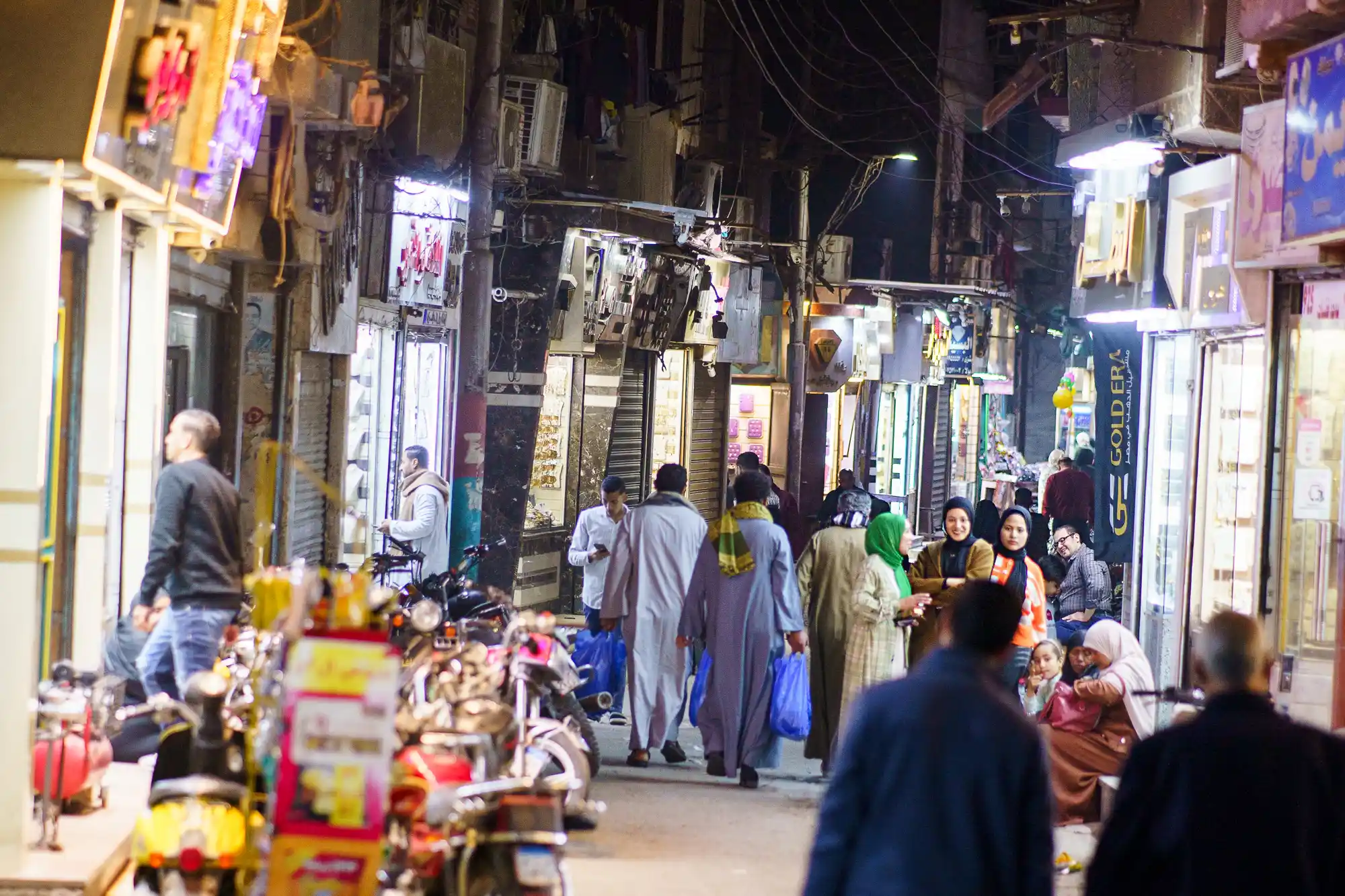 People walk through a narrow, brightly lit passage in Luxor at night, past a row of parked motorcycles.