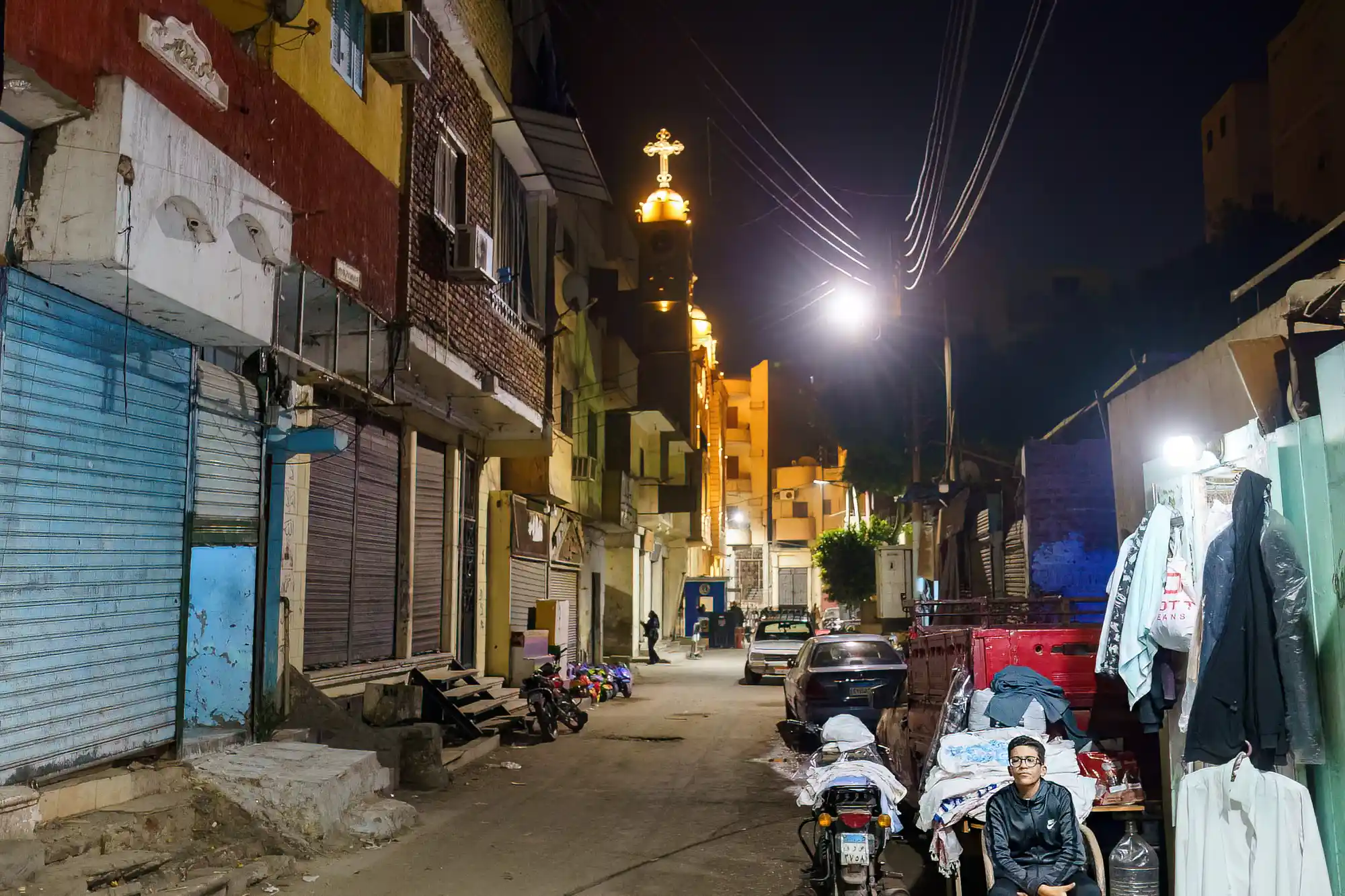 A Luxor street at night with a person in a black jacket passing blue shutters, and warm light glowing from a tower.