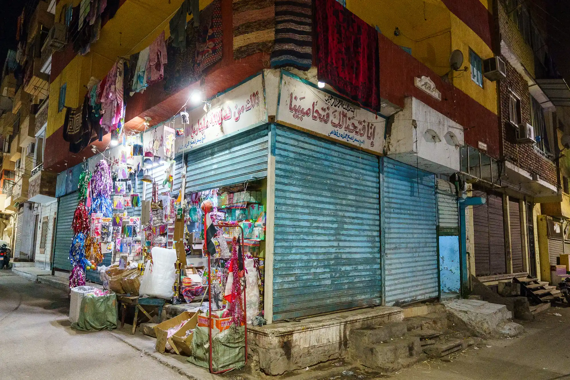 A corner shop with blue metal shutters under bright lights in Luxor, with colorful fabrics hanging above and boxes below.