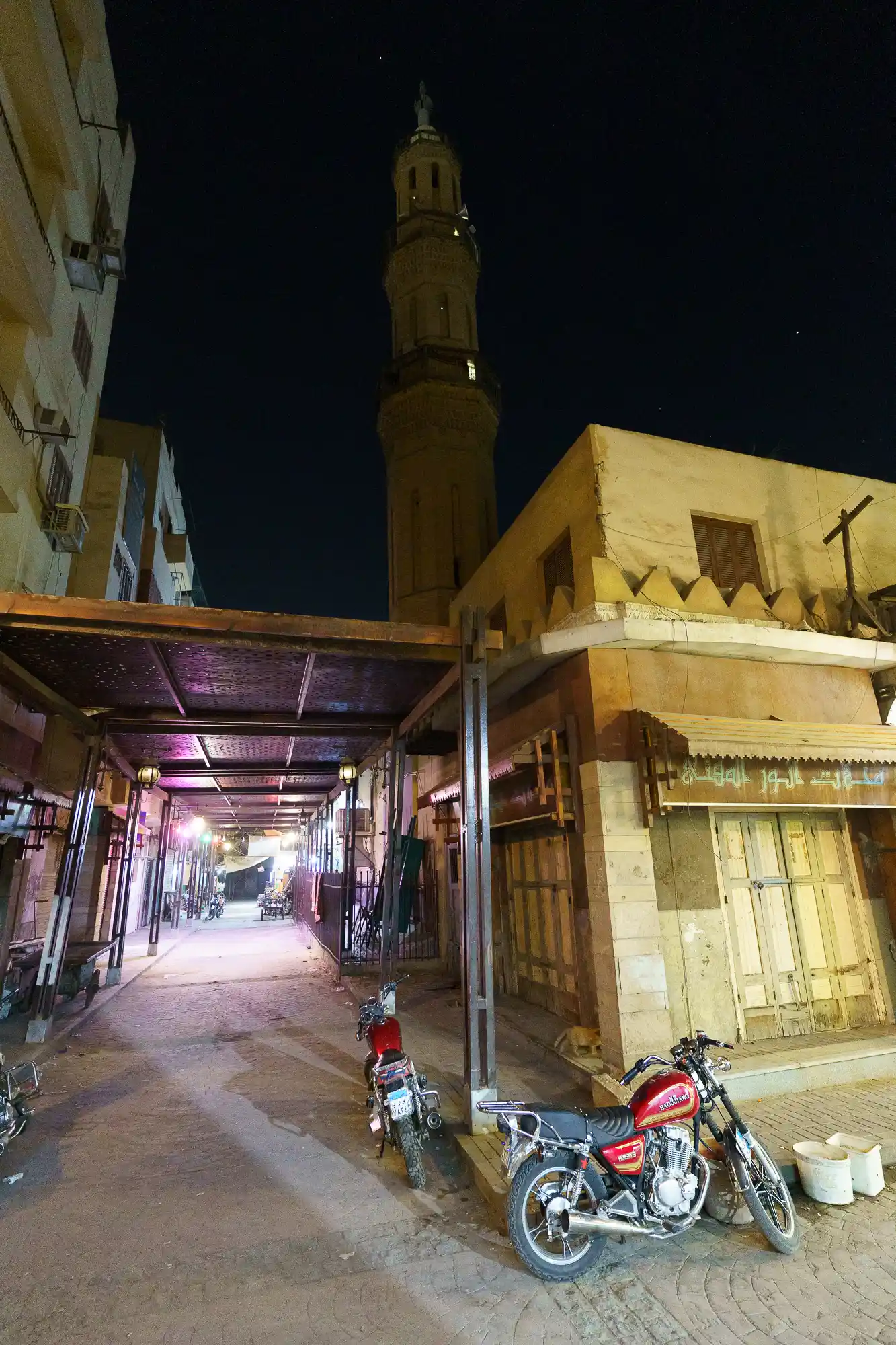 A tall tower rises above a covered walkway in Luxor at night, with red motorcycles parked on stone ground under purple light.
