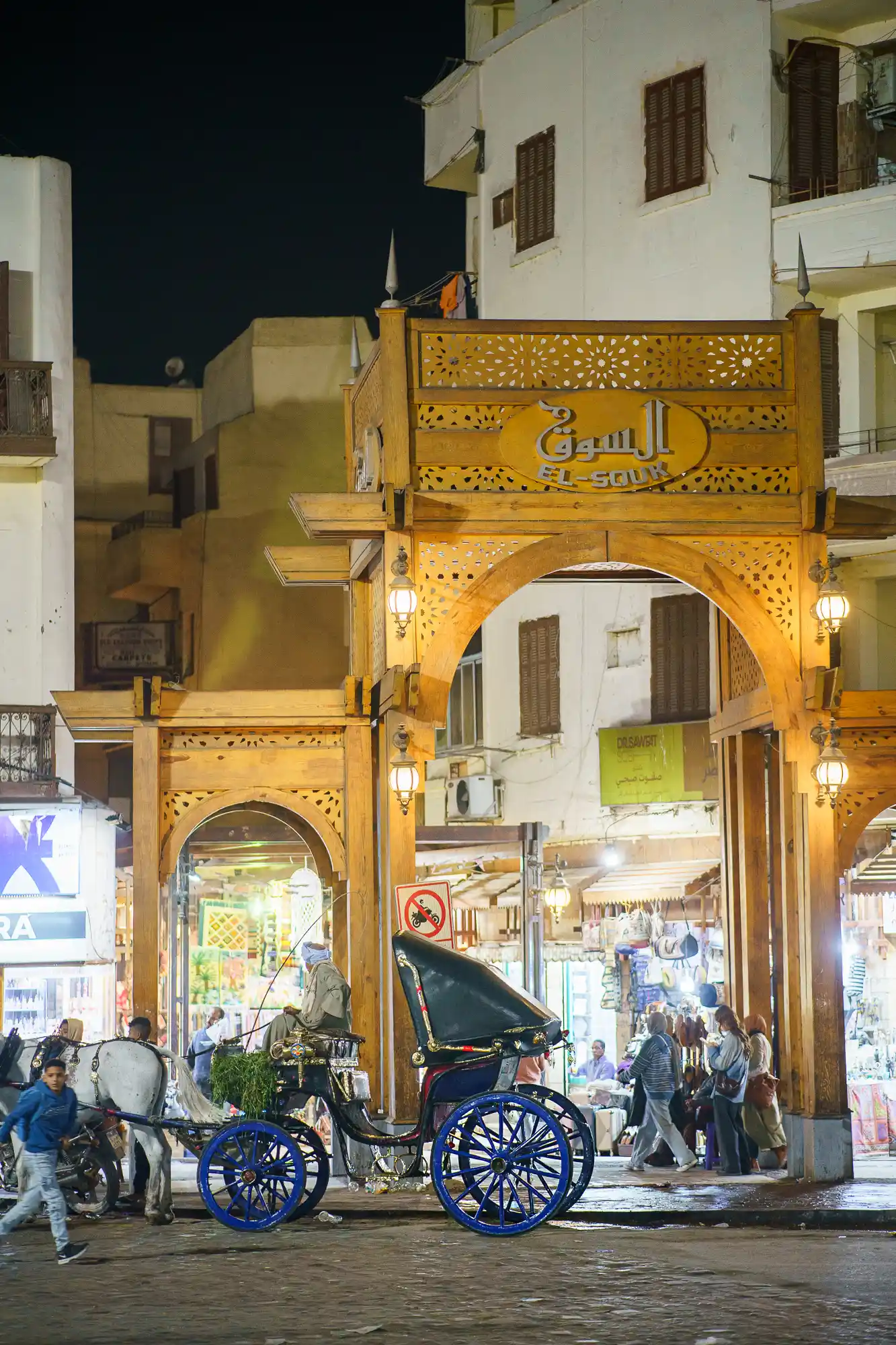 A horse carriage waits under a carved wooden arch in Luxor at night, as people walk past in the warm light.