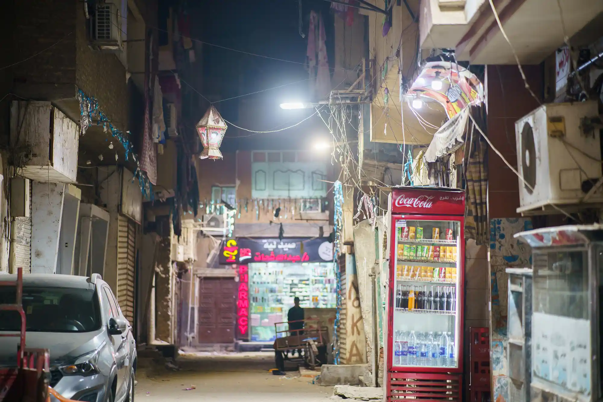 A narrow Luxor alley at night, lit by a hanging lantern and a red glass cooler, with a silver car in the shadows.