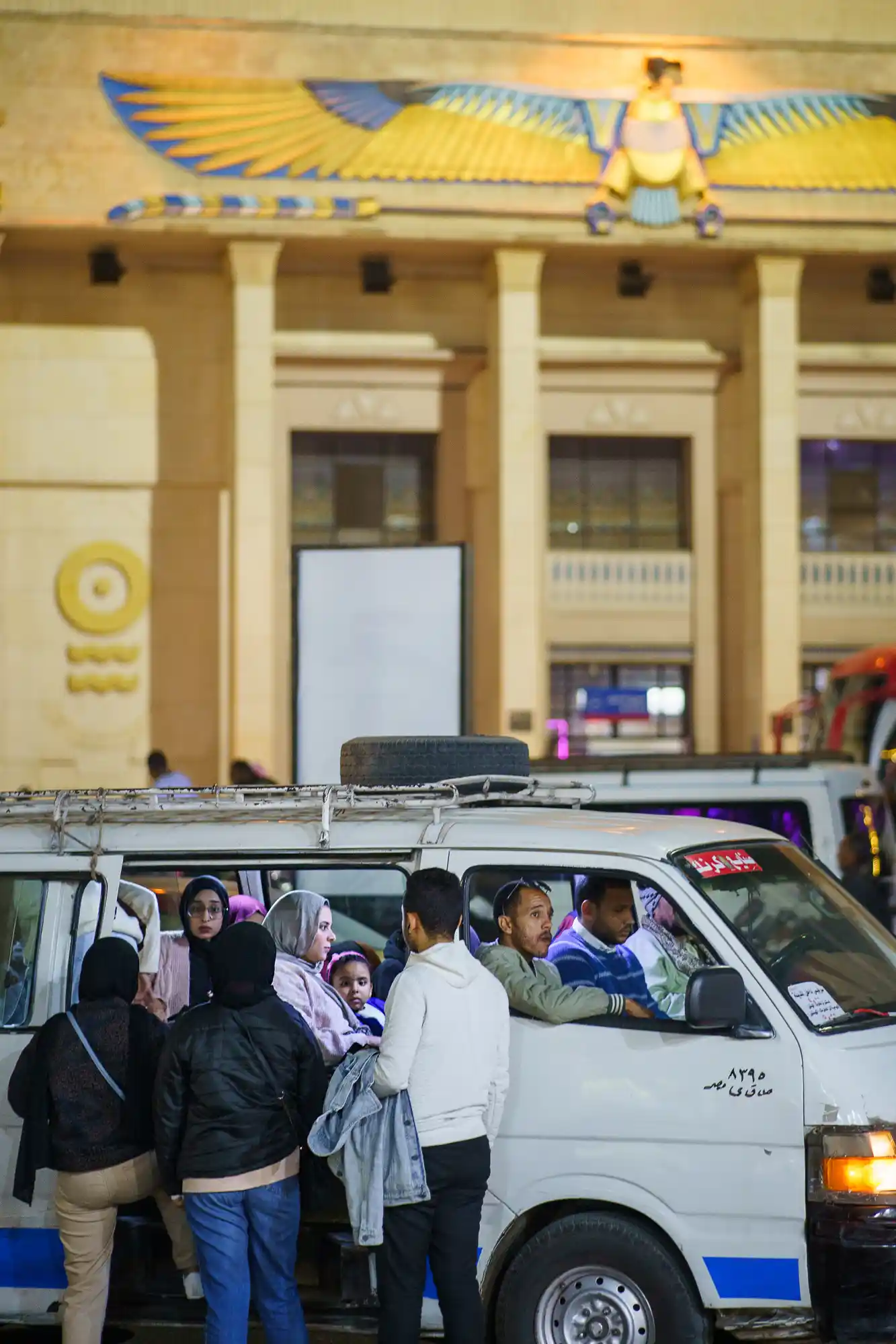 People board a white van with a spare tire on the roof in Luxor at night, under a glowing yellow and blue winged sign.