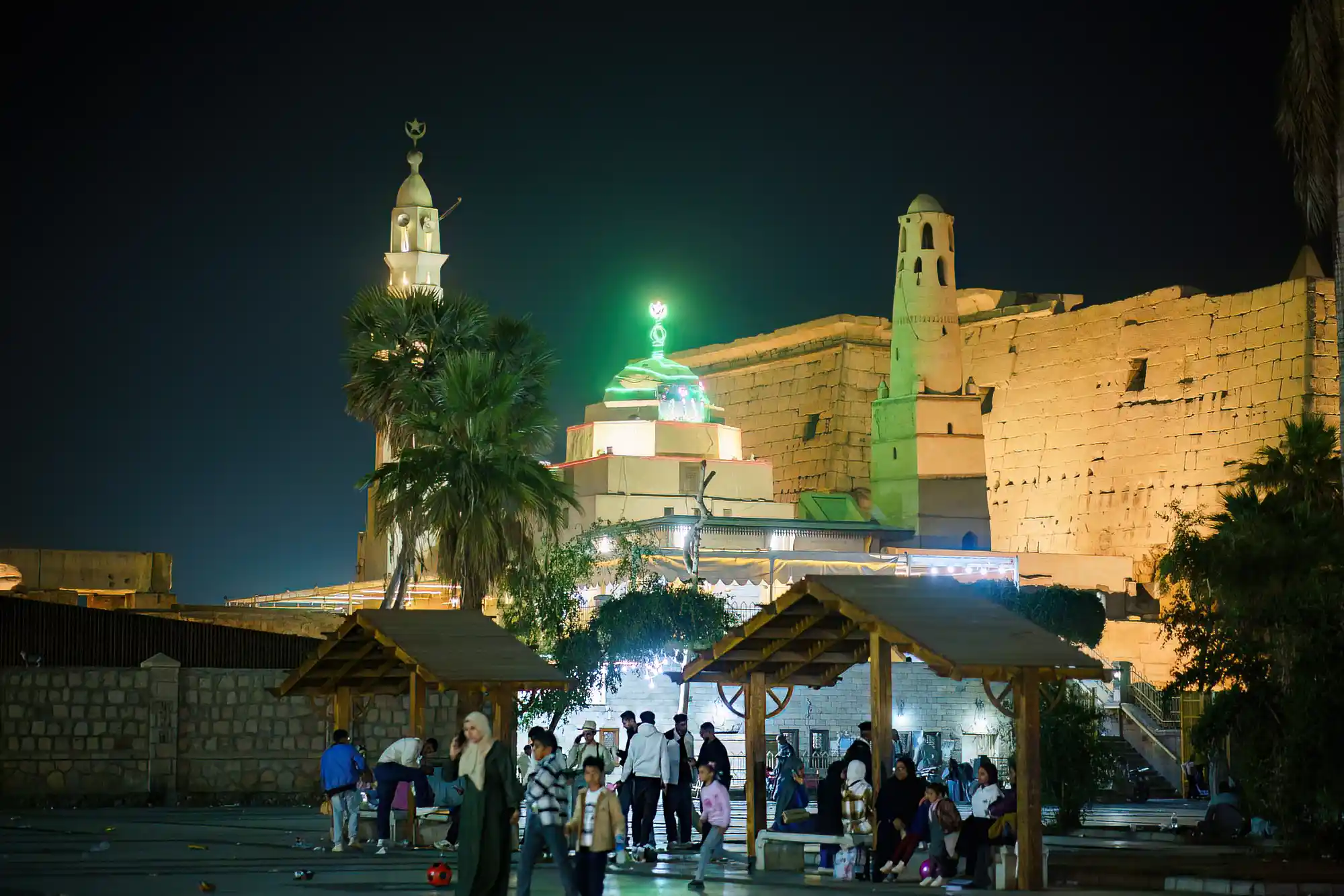 People gather by wooden shelters in warm light in Luxor at night, with a stone structure glowing green behind them.
