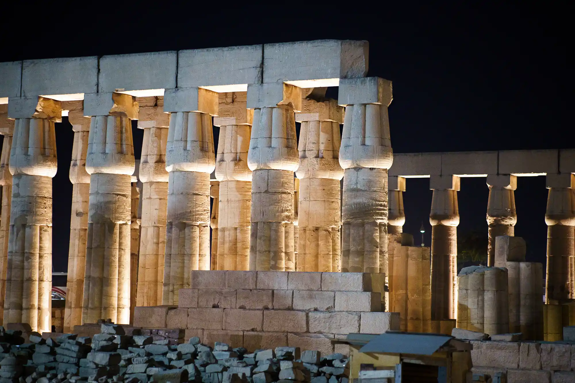 Stone columns lit from below at night in Luxor, glowing warm yellow above dark rubble in the foreground.