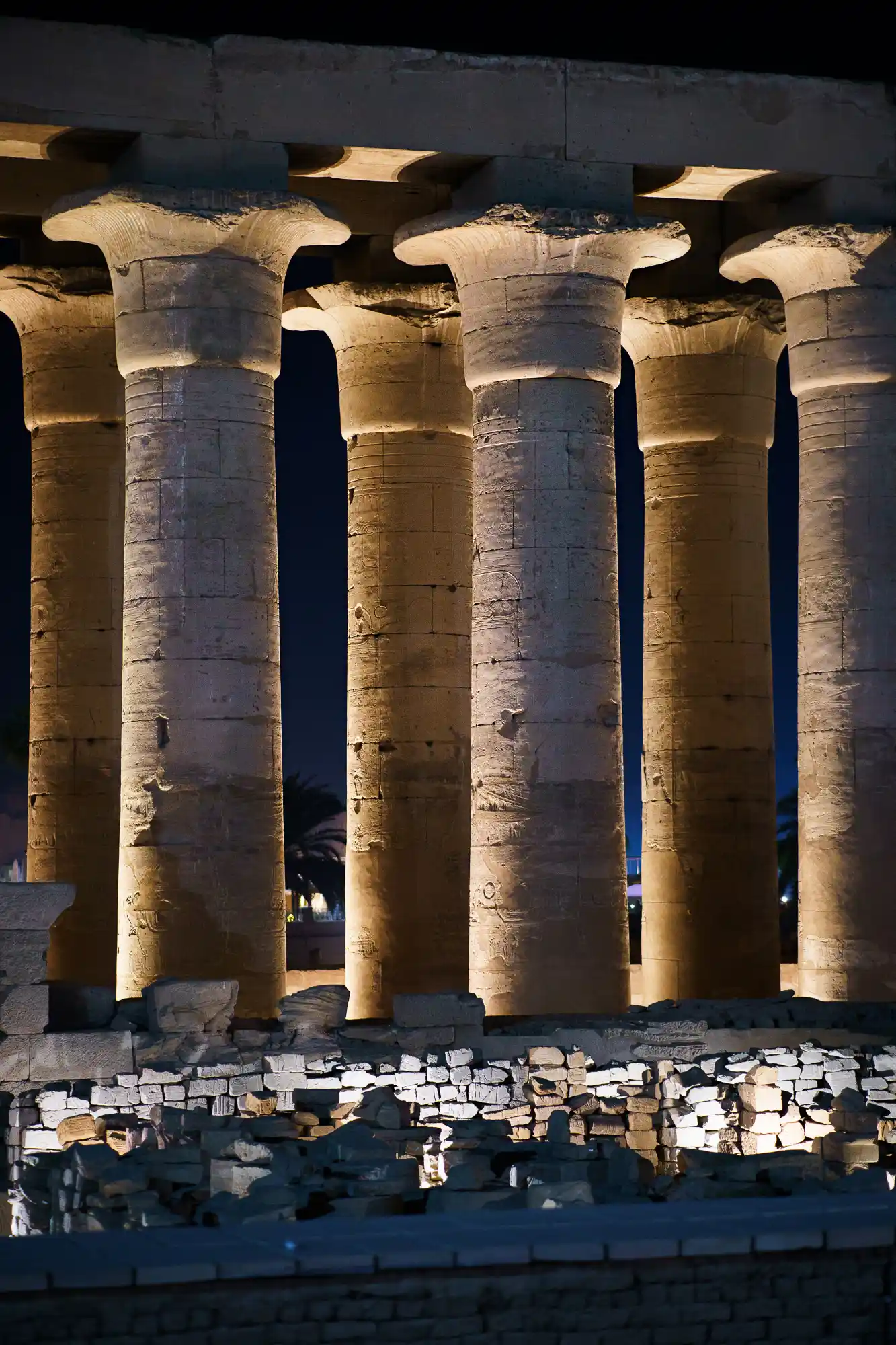 Floodlit stone columns in Luxor at night, warm yellow light on the pale stone above rough blocks in the foreground.