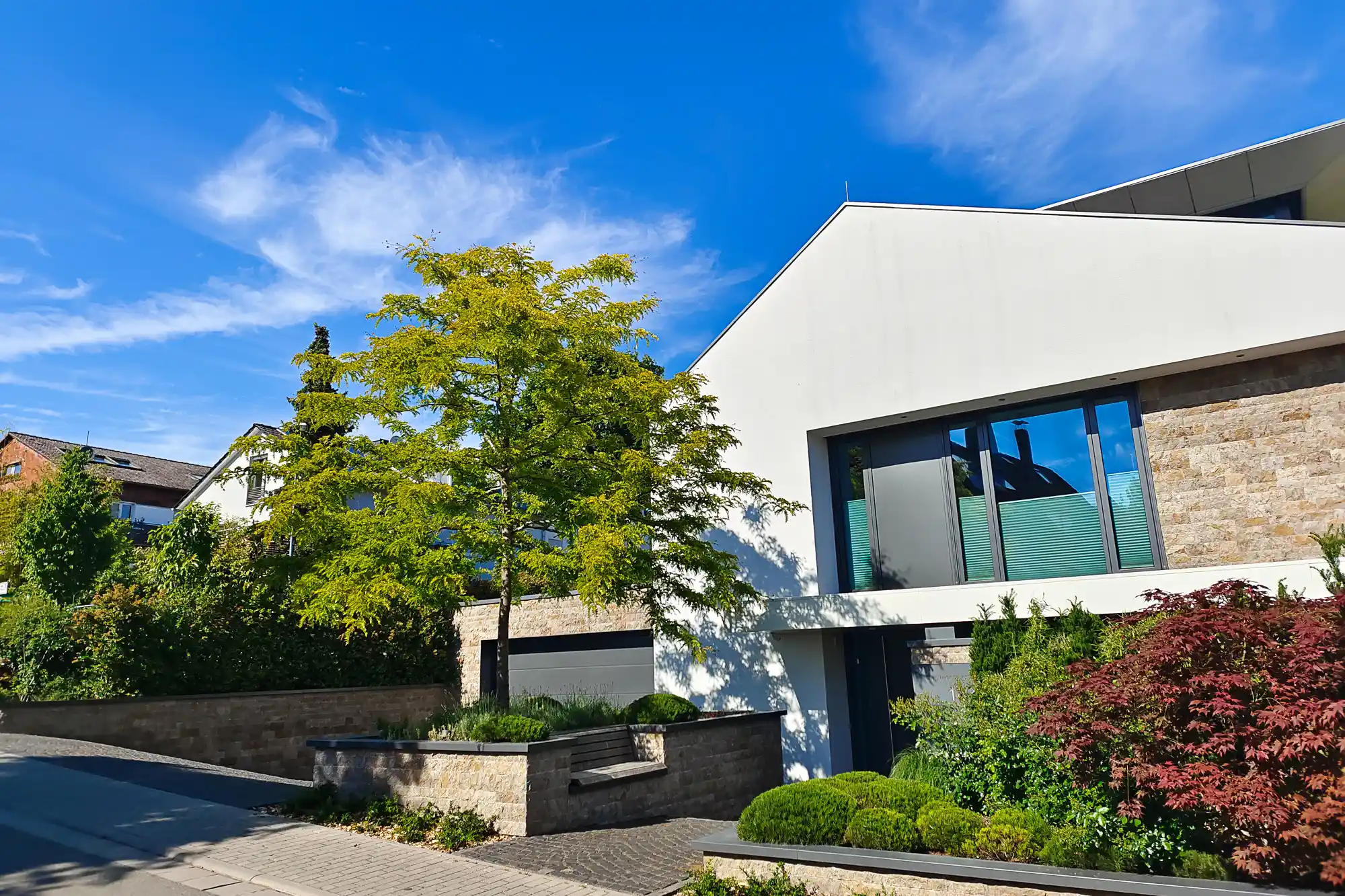 A white building with stone sections and a green tree in Traisa, with red bushes in bright sunlight.