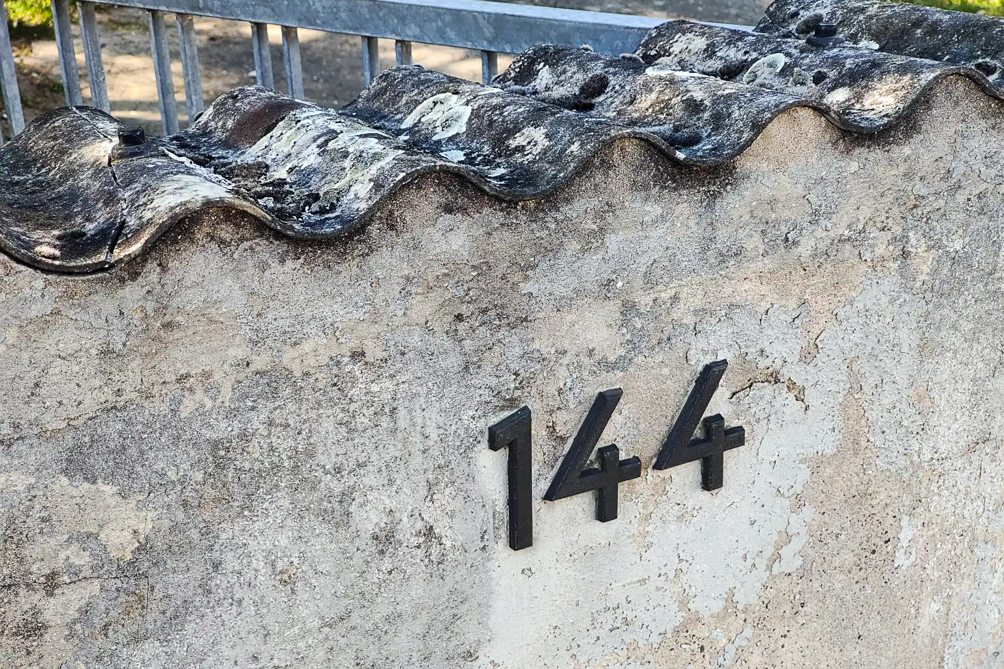Black numbers on a light stone wall beneath dark wavy roof tiles in Traisa, with bright light on the rough surface.