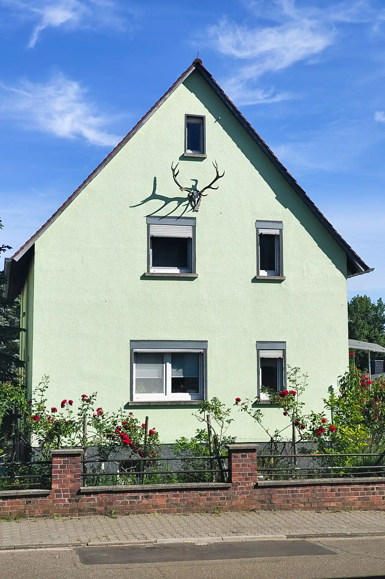 A light green house in Traisa with a deer sculpture and red flowers, sunlight casting shadows on the textured wall.