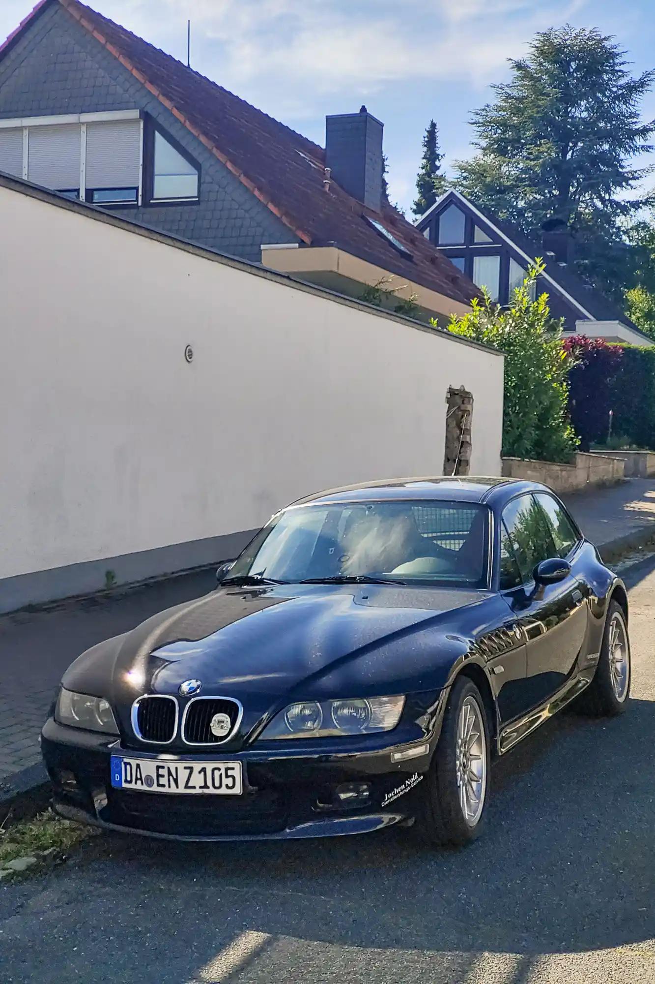 A shiny black car with silver wheels parked in Traisa, sunlight reflecting off its body near a white wall.