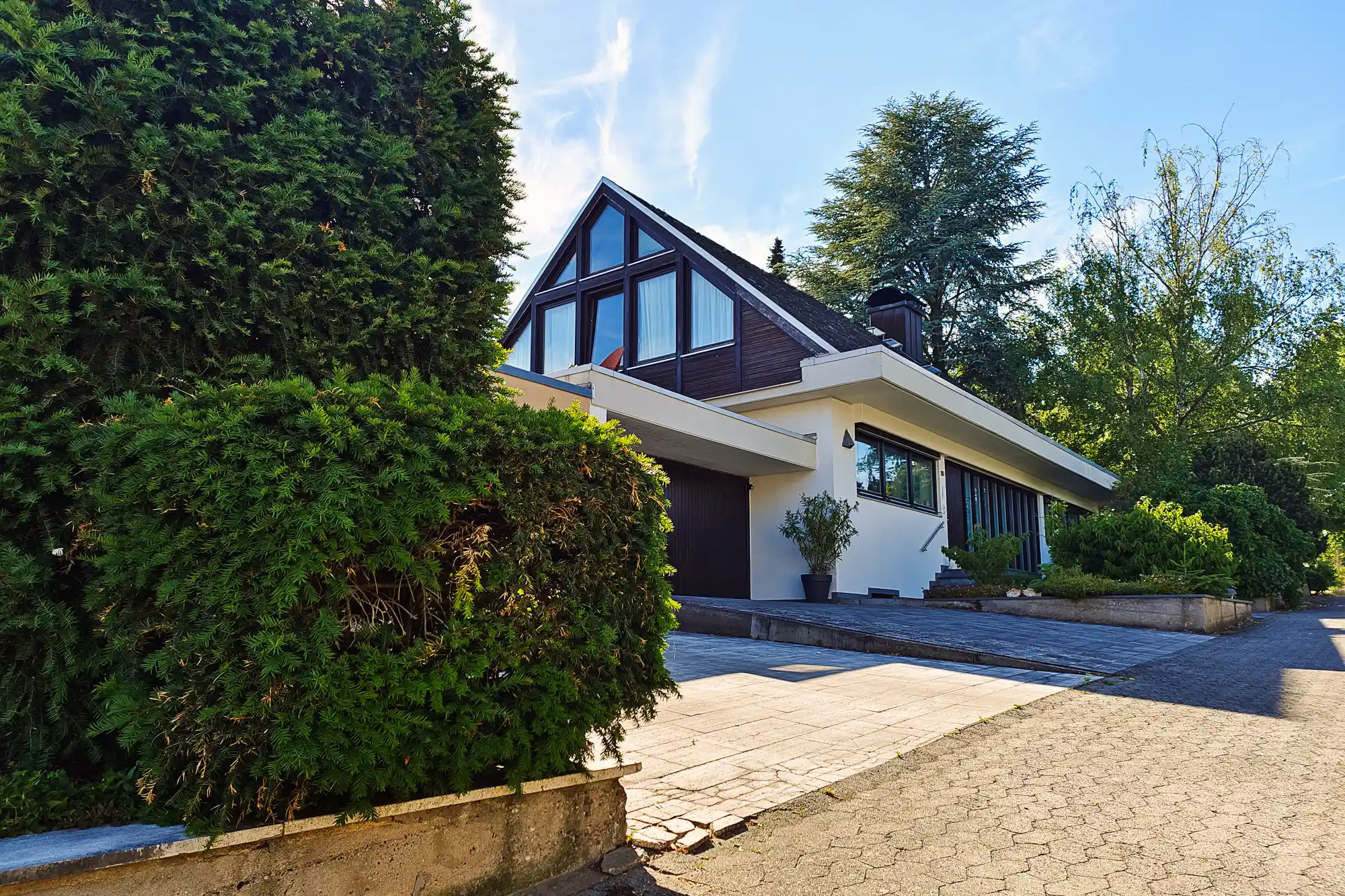 A white and dark house with large windows in Traisa, with green foliage and bright light on the stone path.