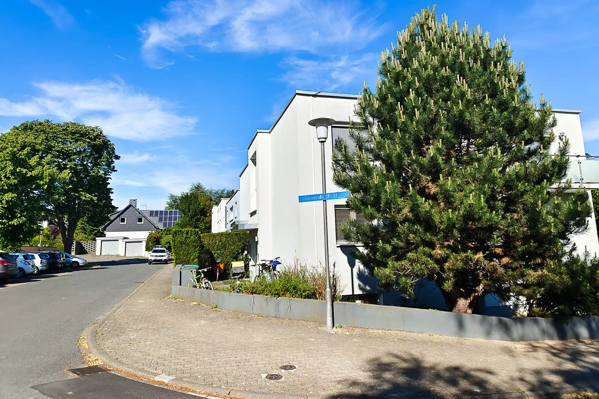 White buildings in Traisa under a blue sky with wispy clouds, a tall green tree casting shadows on the paved ground.