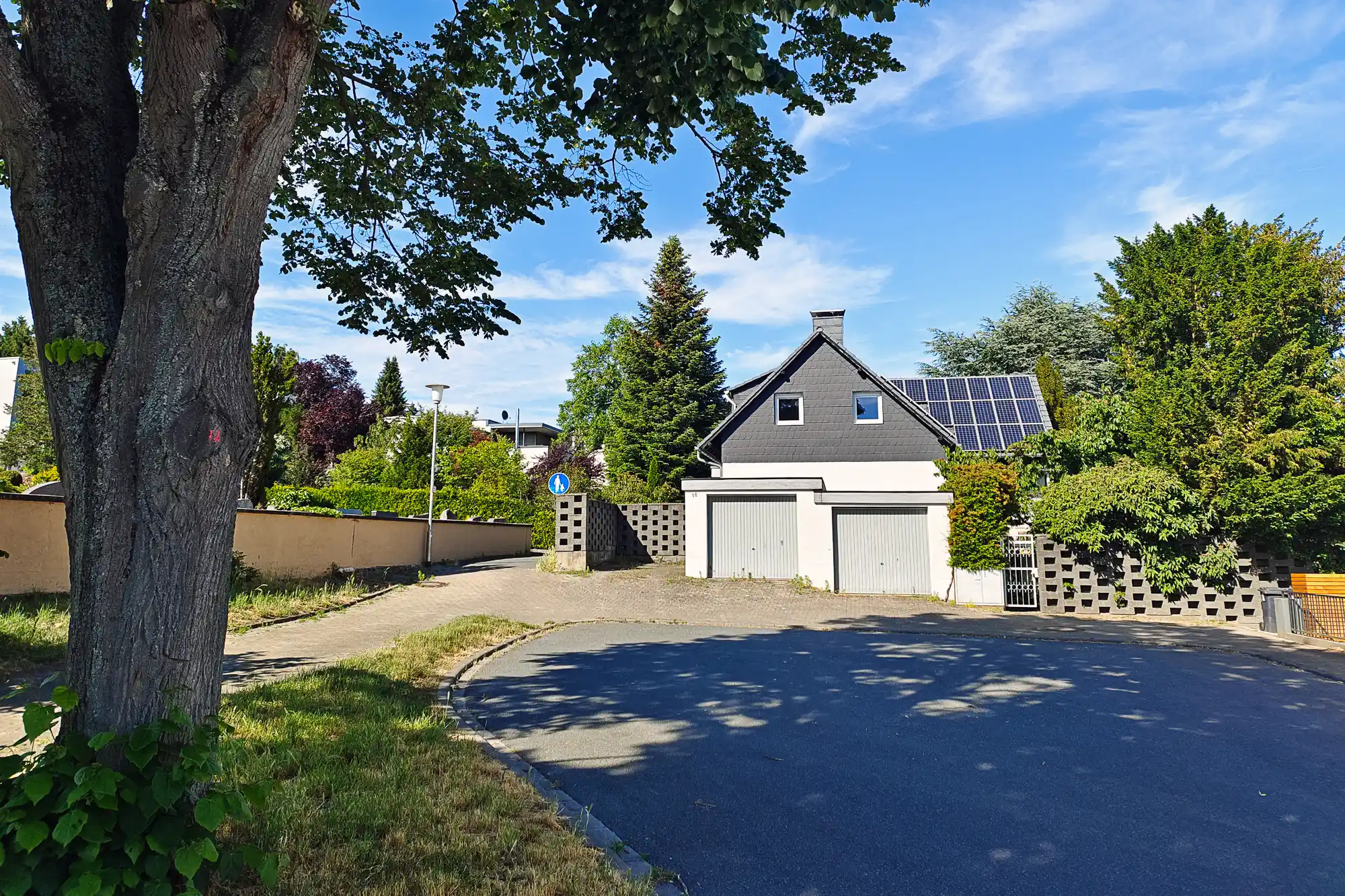 A house with a dark roof and solar panels behind a tree in Traisa, sunlight casting shadows on the paved road.