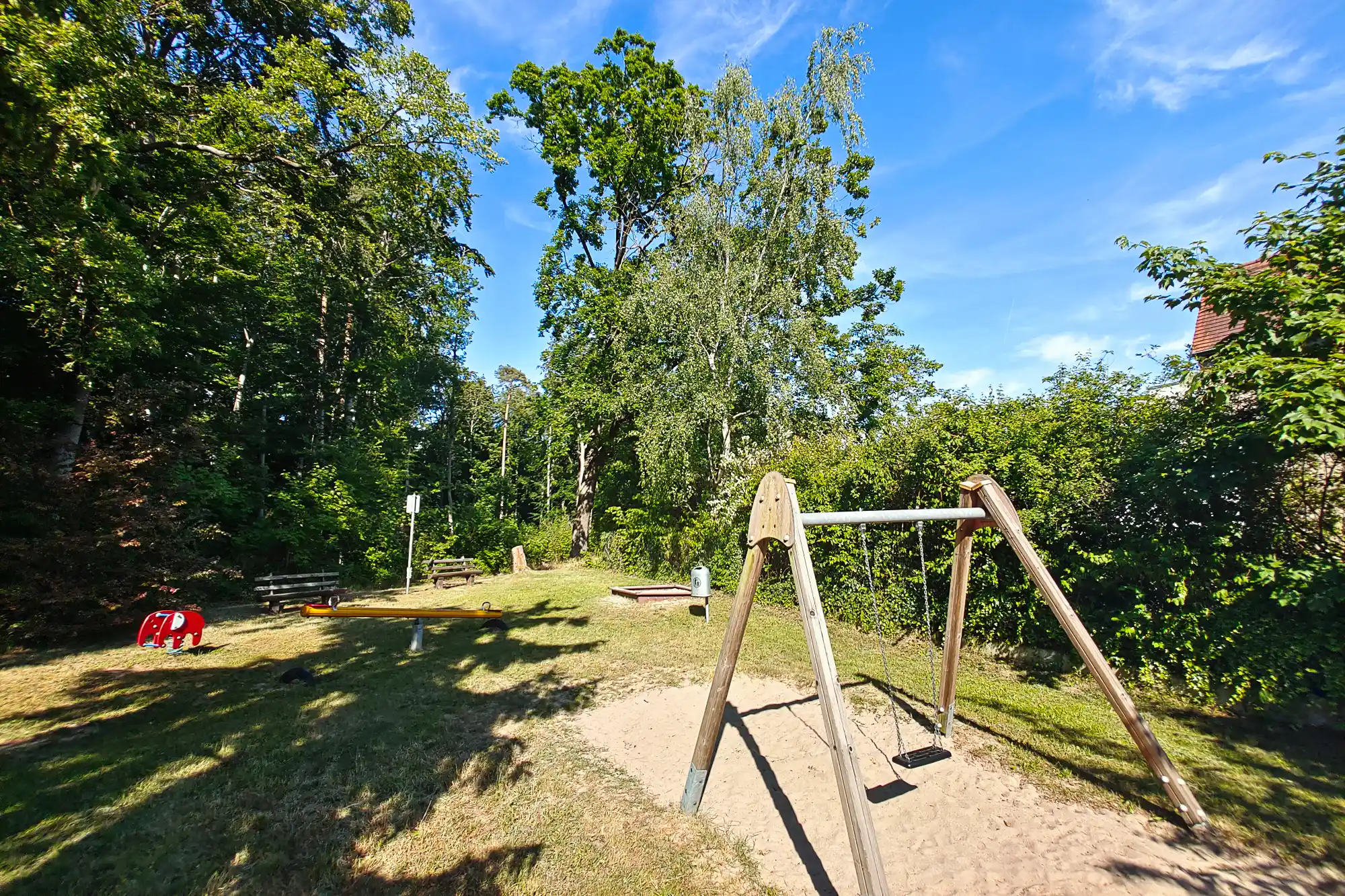 A wooden swing set in the sand at a playground in Traisa, with a red toy and green trees casting long shadows.