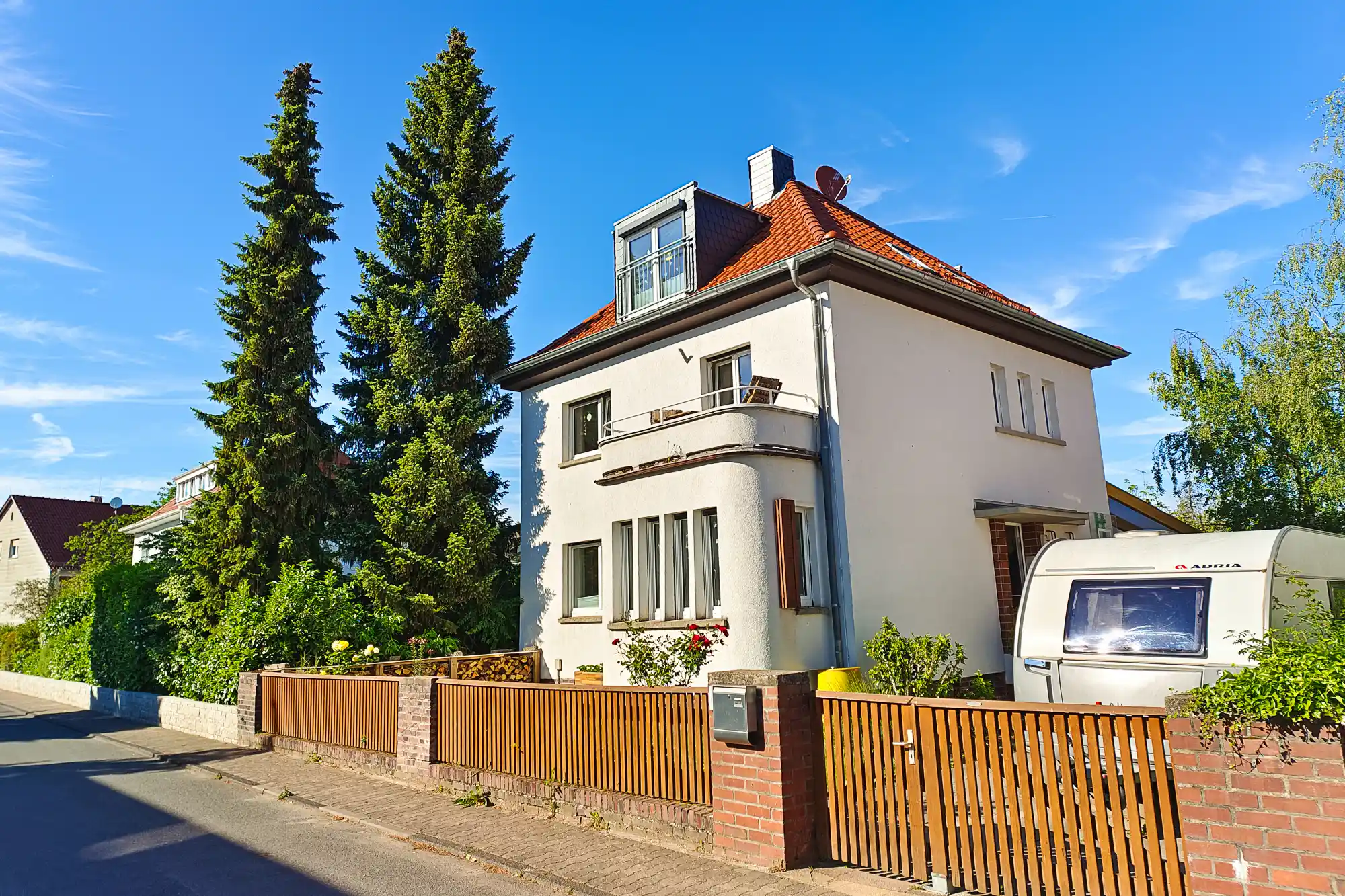A white house with a red tile roof in Traisa, with two tall green trees and a white trailer behind a wooden fence.