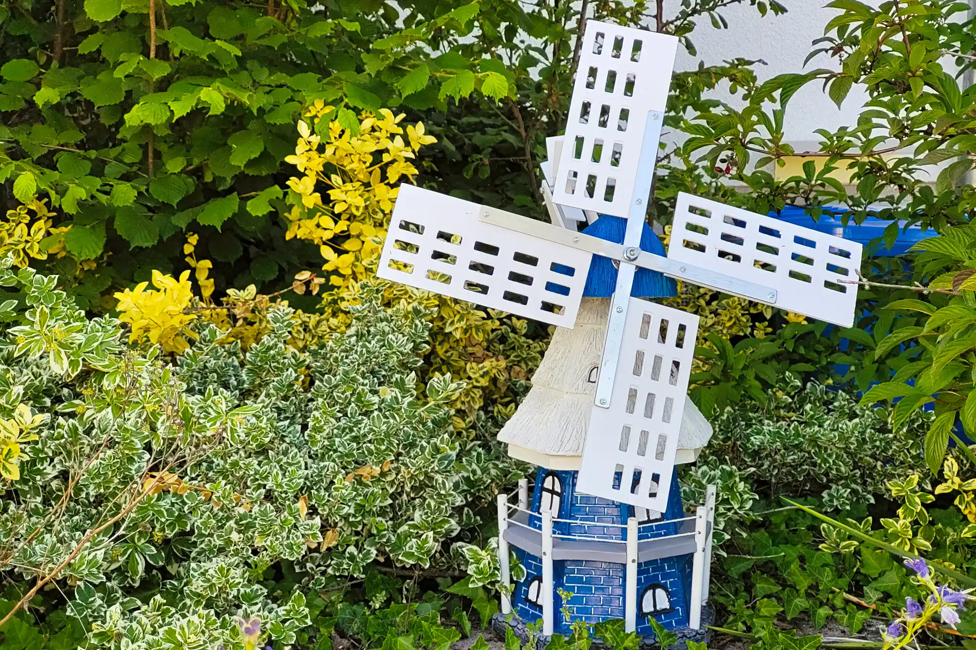 A blue and white model windmill with lattice blades and a textured roof among green and yellow leaves in Traisa.