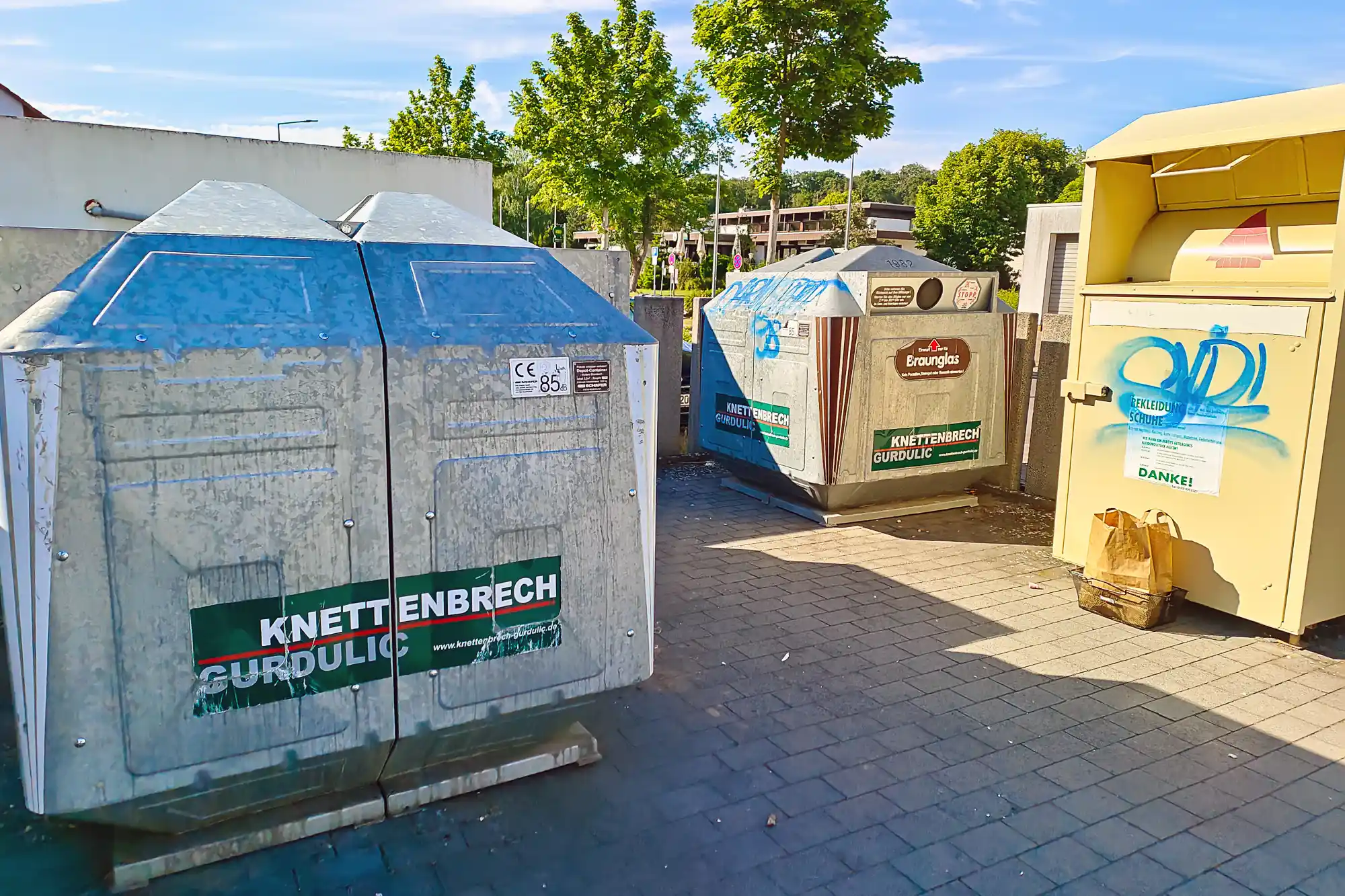 Metal bins with blue tops and green labels on brick paving in Traisa, sunlight casting sharp shadows.