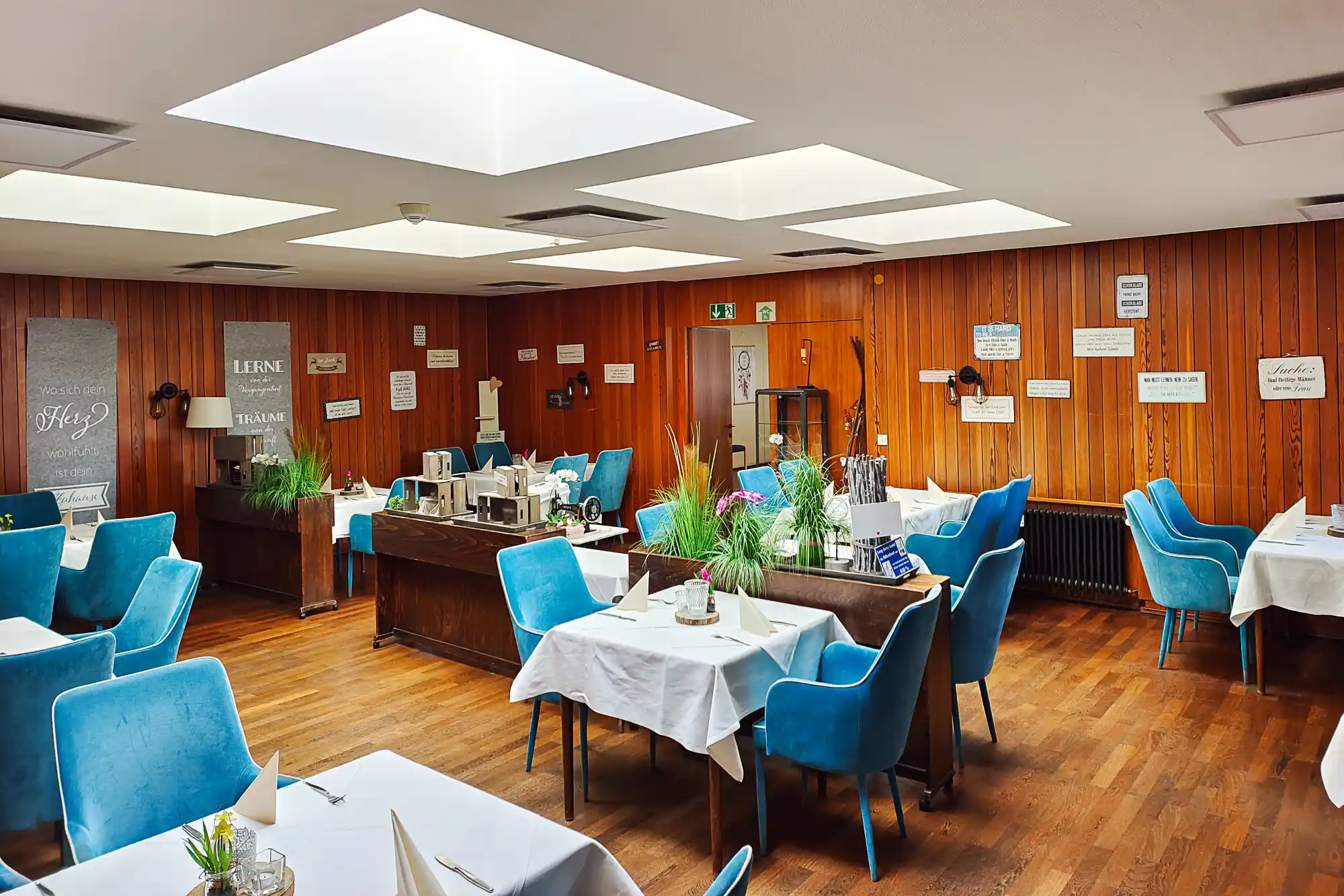 The dining room of the Bürgerhaus in Traisa, with blue chairs, wooden walls and white tablecloths under bright overhead light.